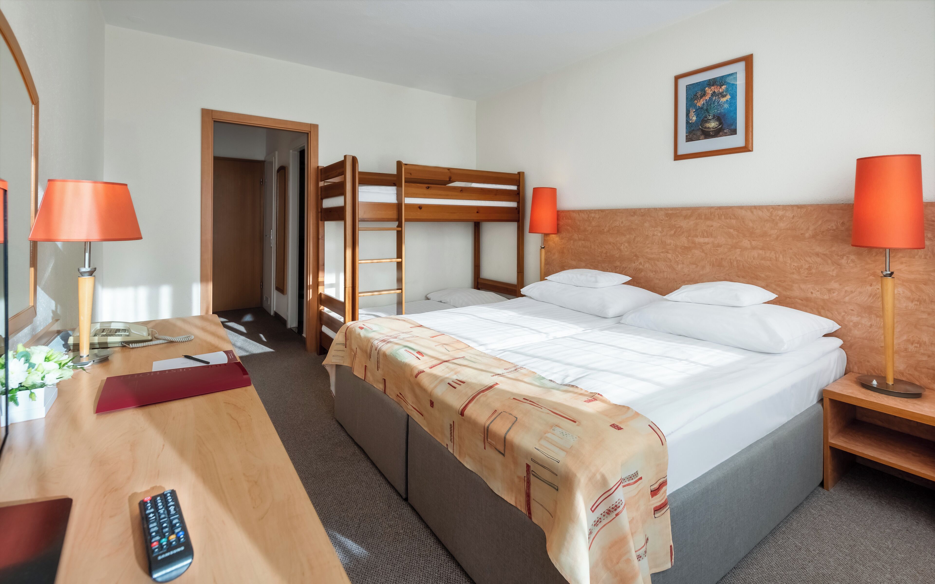 A hotel room with a bunk bed, a desk with a telephone, a remote control, and two lamps.