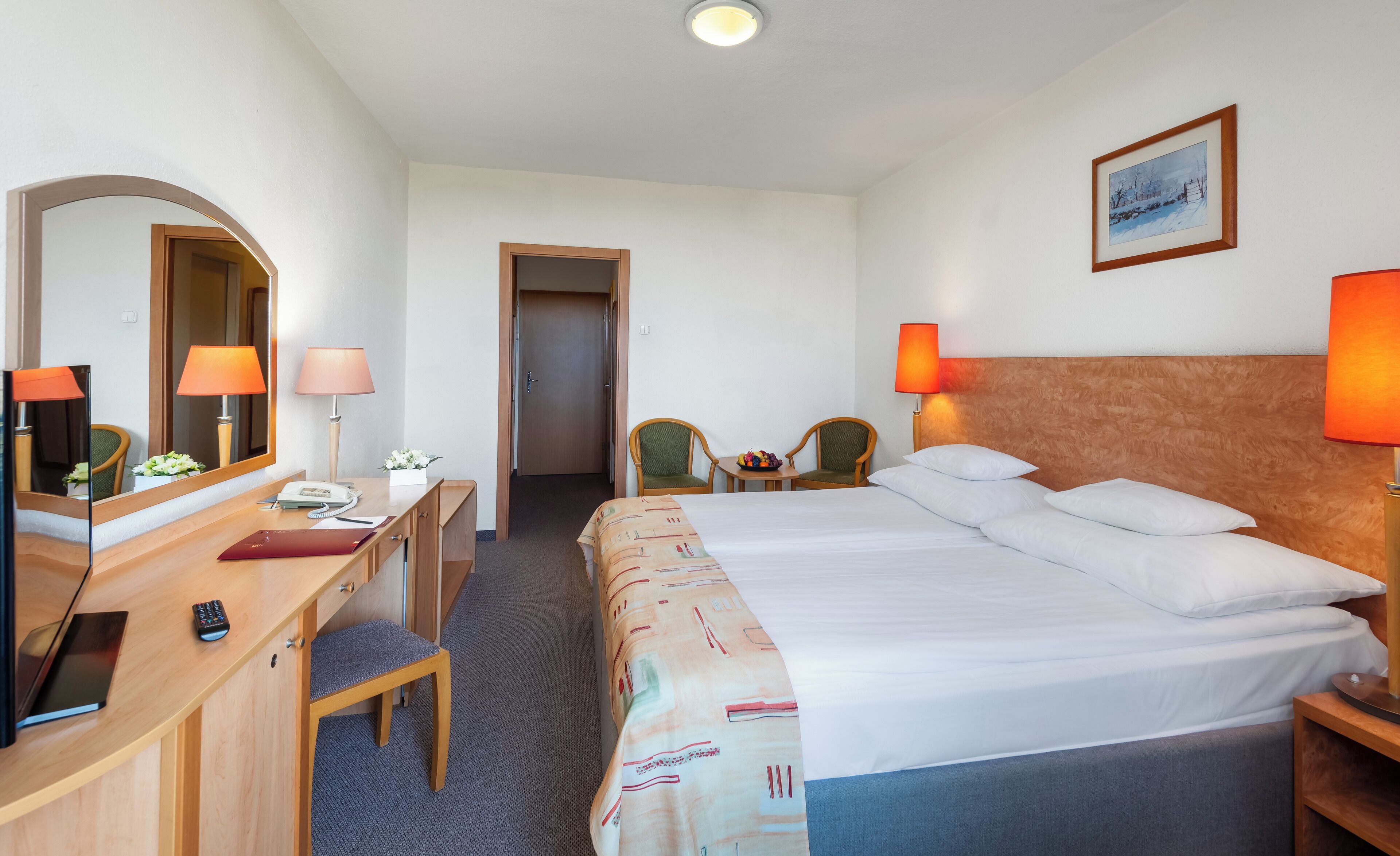 Una habitación de hotel con una cama, dos sillas, un escritorio con lámpara, un espejo y un cuadro en la pared.