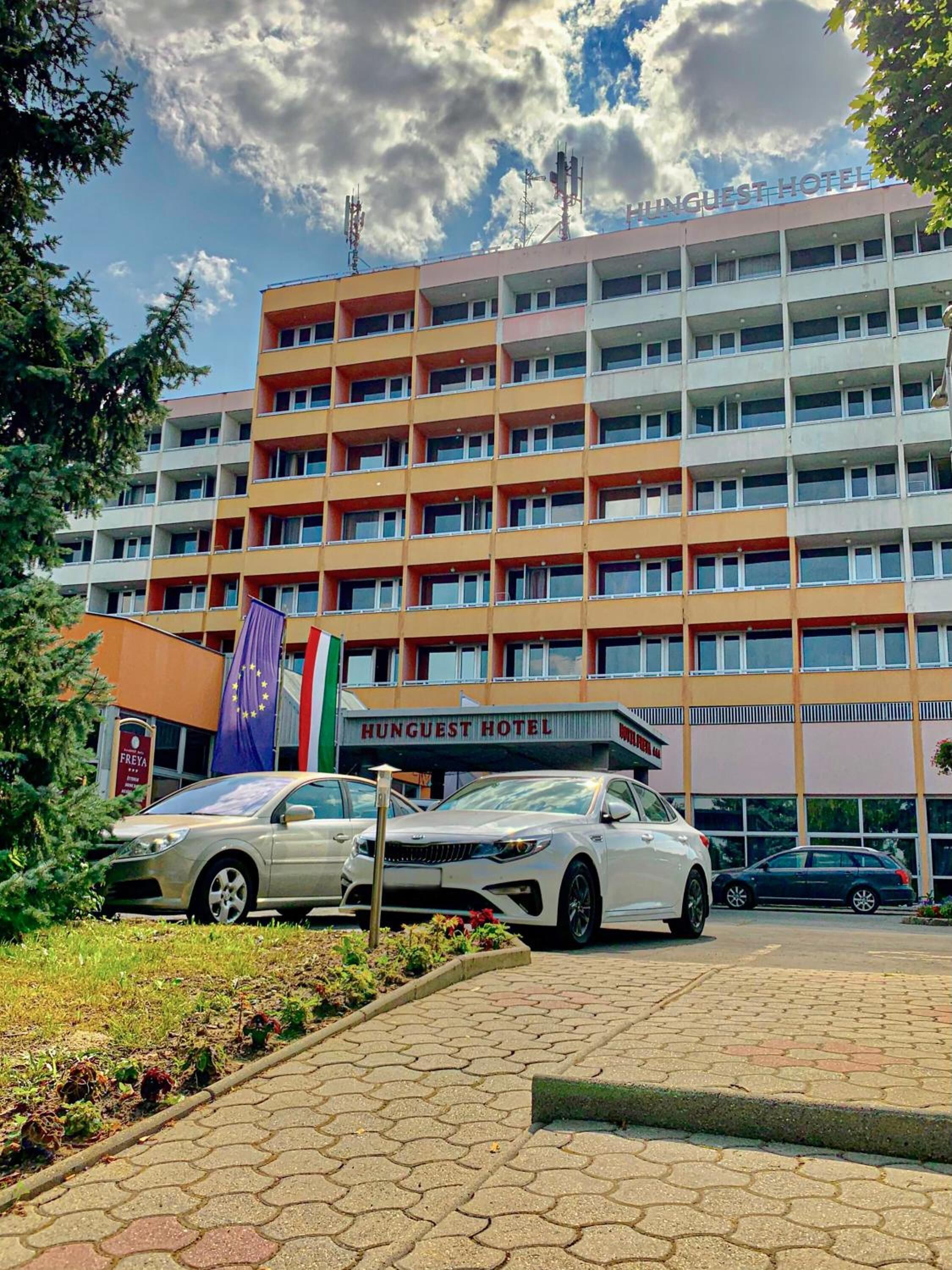 A multi-story building with the sign "HUNGUEST HOTEL" prominently displayed. There are several cars parked in front, and a flag with a star pattern is visible.