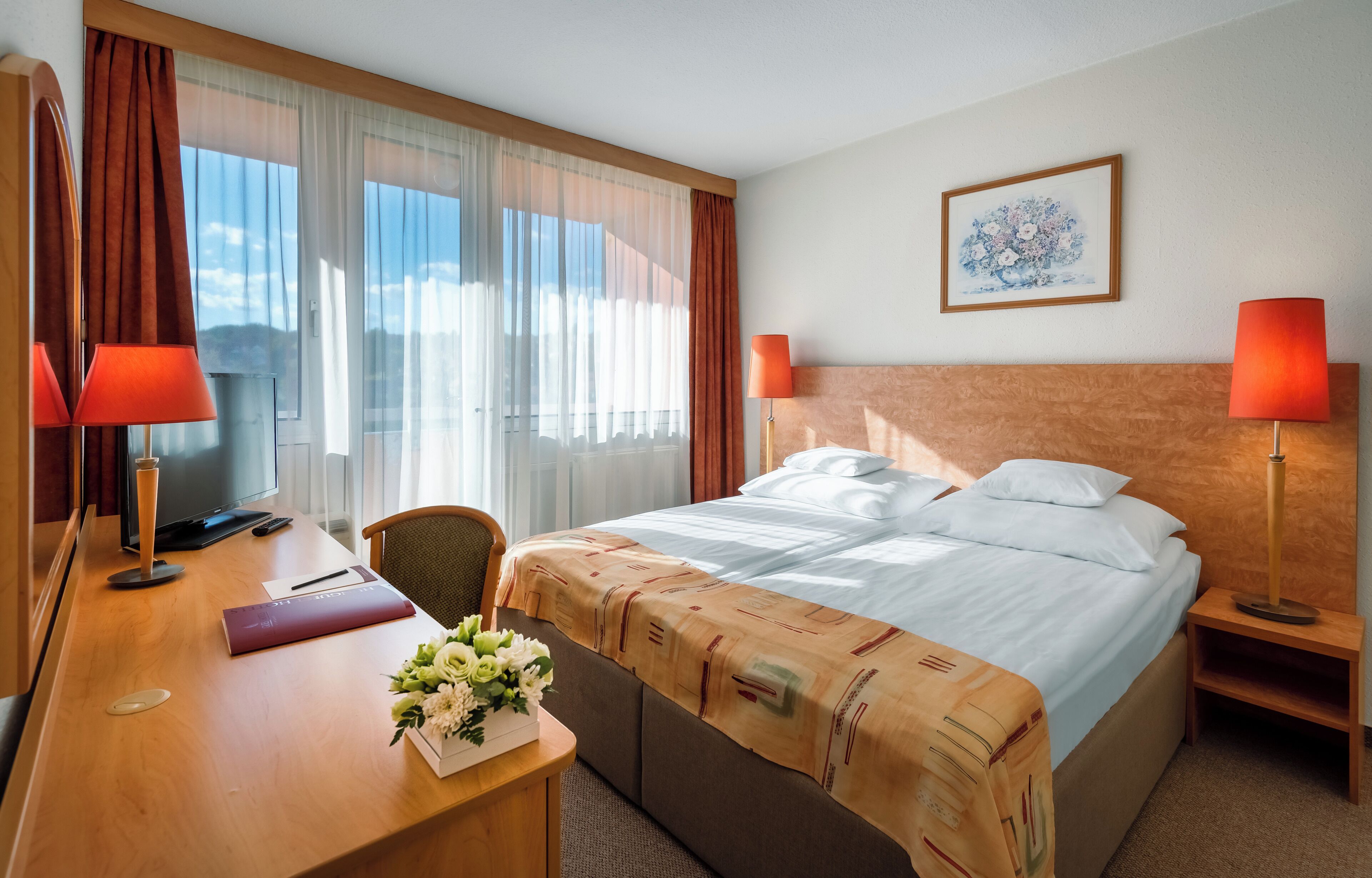 A hotel room with a bed, two bedside lamps, a desk with a computer, a chair, a desk lamp, a vase with flowers, and a framed picture on the wall.