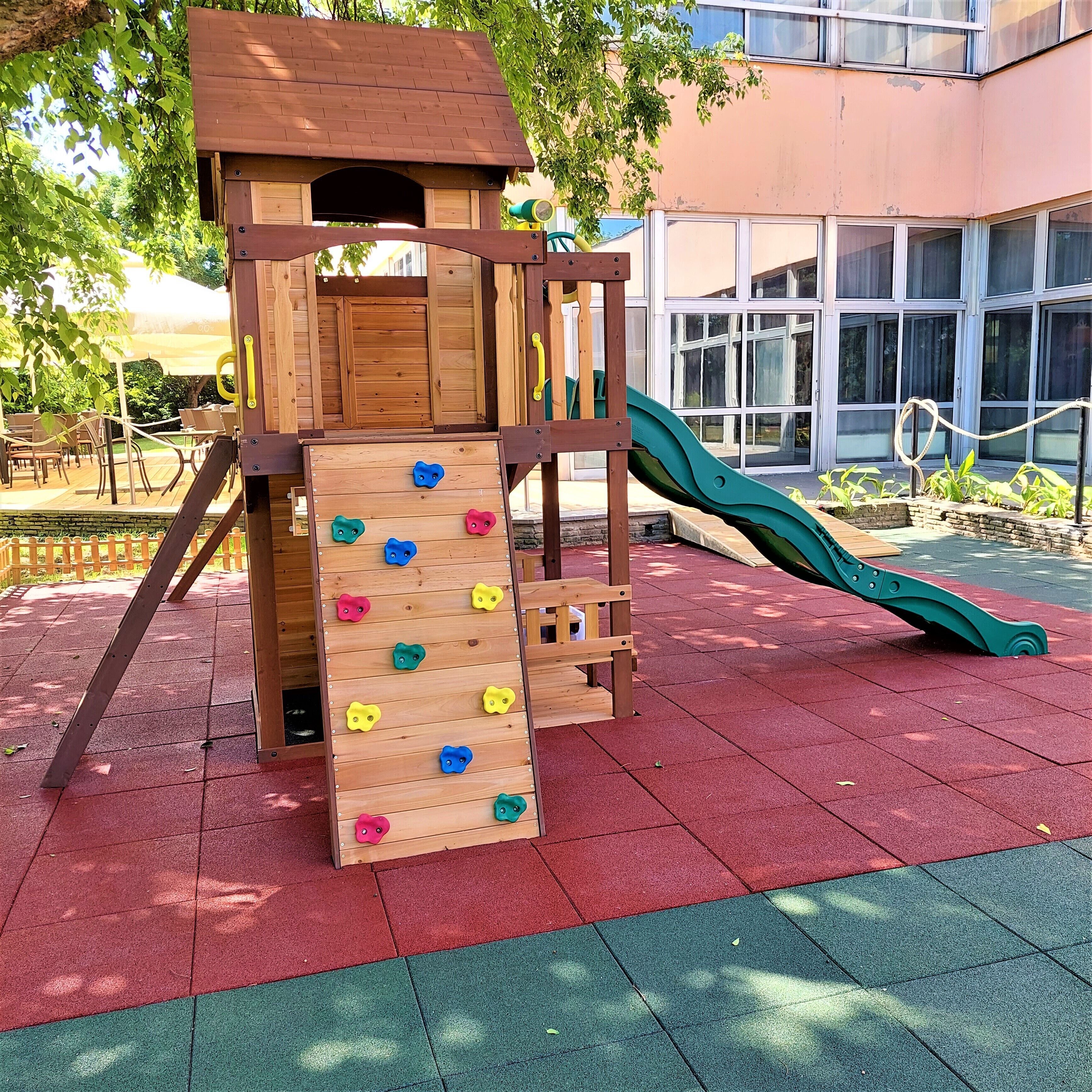 A wooden play structure with a climbing wall and slide, set on red and green rubber flooring, with a building in the background.
