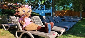 A mascot sitting on a row of lounge chairs in a backyard with a wooden fence and a building in the background. - Hunguest Hotel Freya (Zalakaros)