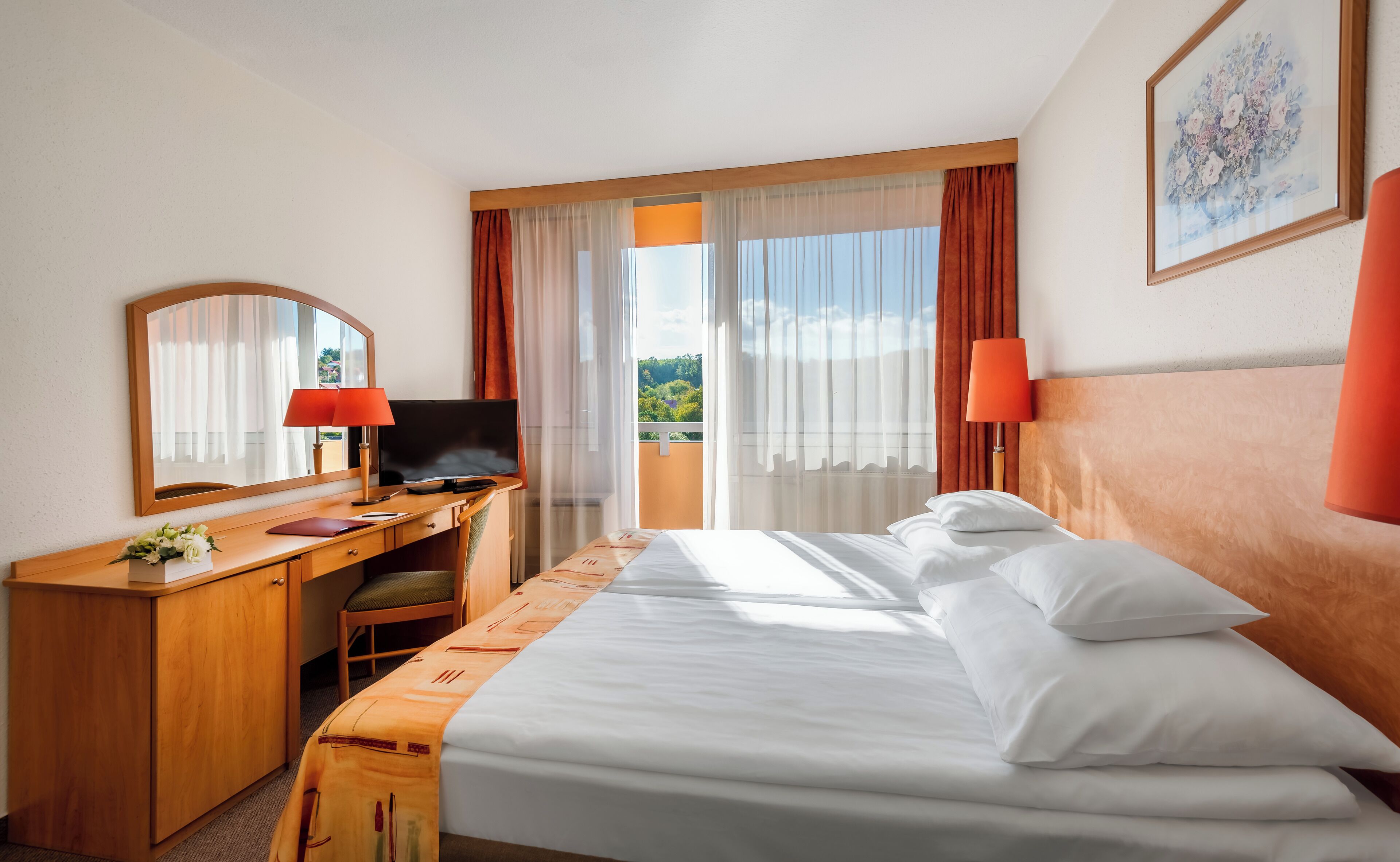 Quarto de hotel com cama, escrivaninha, cadeira, televisão e vista para o verde lá fora.