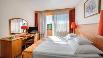Quarto de hotel com cama, escrivaninha, cadeira, televisão e vista para o verde lá fora.