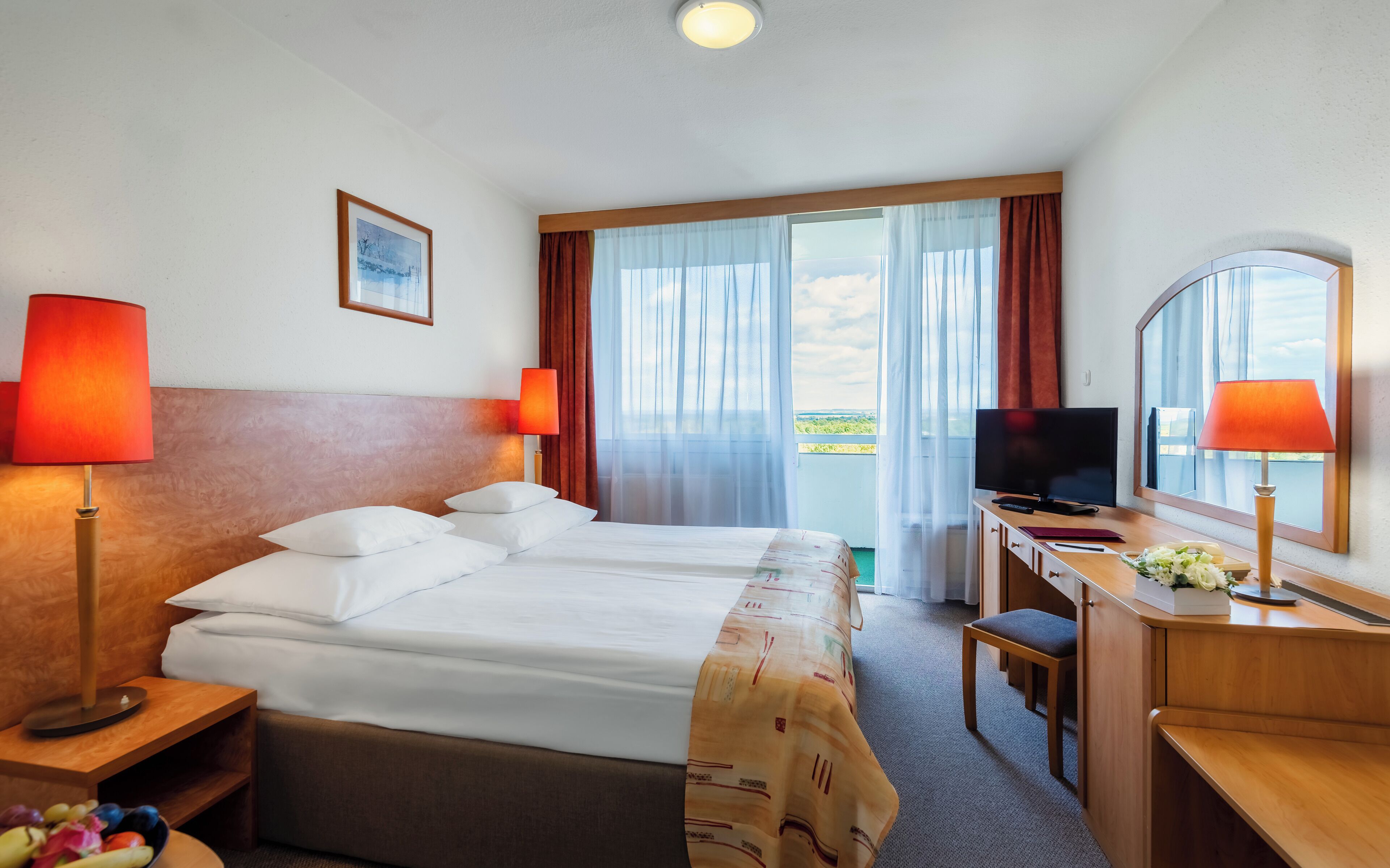 Habitación de hotel con una cama grande, dos lámparas de noche, un escritorio con televisor y vistas al exterior a través de un ventanal.