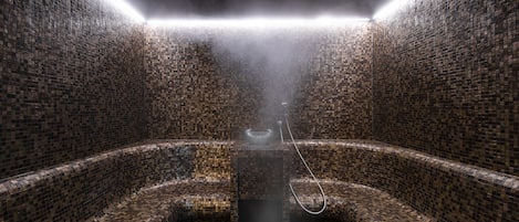 A steam room with mosaic tile walls and benches, a central steam vent, and a showerhead.