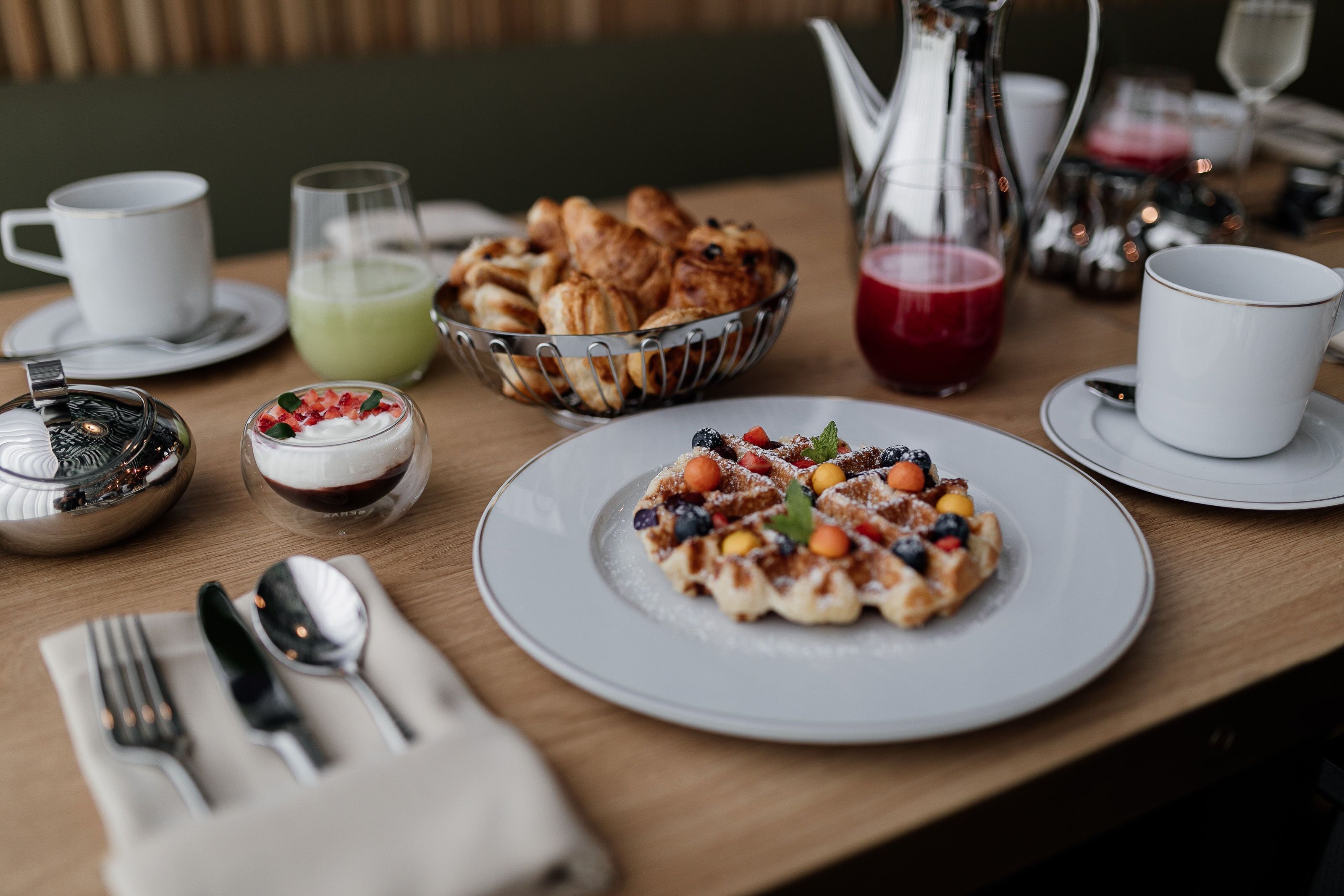 Une table dressée pour le petit-déjeuner, avec une assiette de gaufres garnies de fruits, un bol de croissants et diverses boissons.