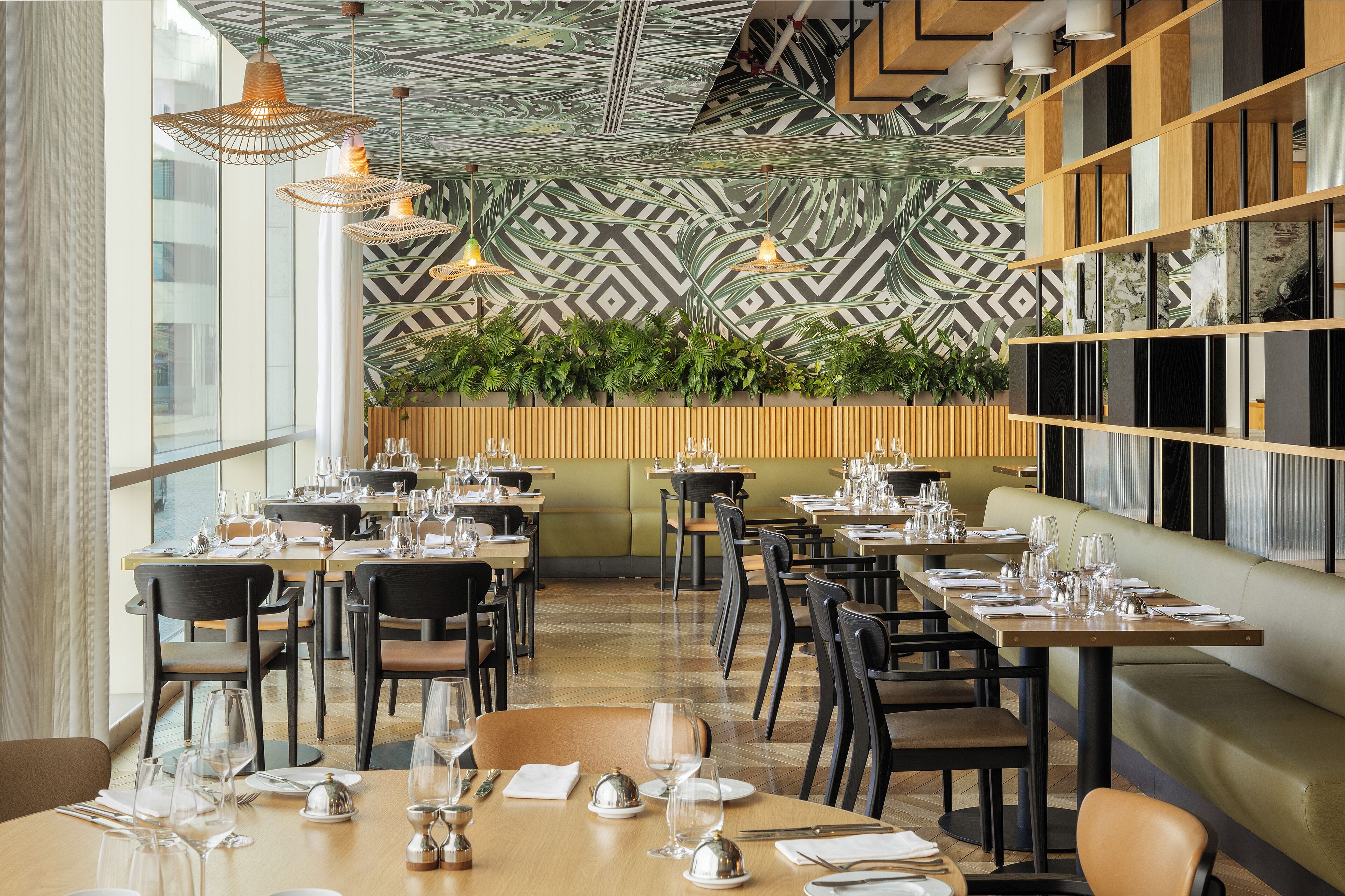 A restaurant with a modern interior, featuring a patterned wall, wooden paneling, and hanging pendant lights. Tables are set with glassware and plates, and there are plants on the wall.