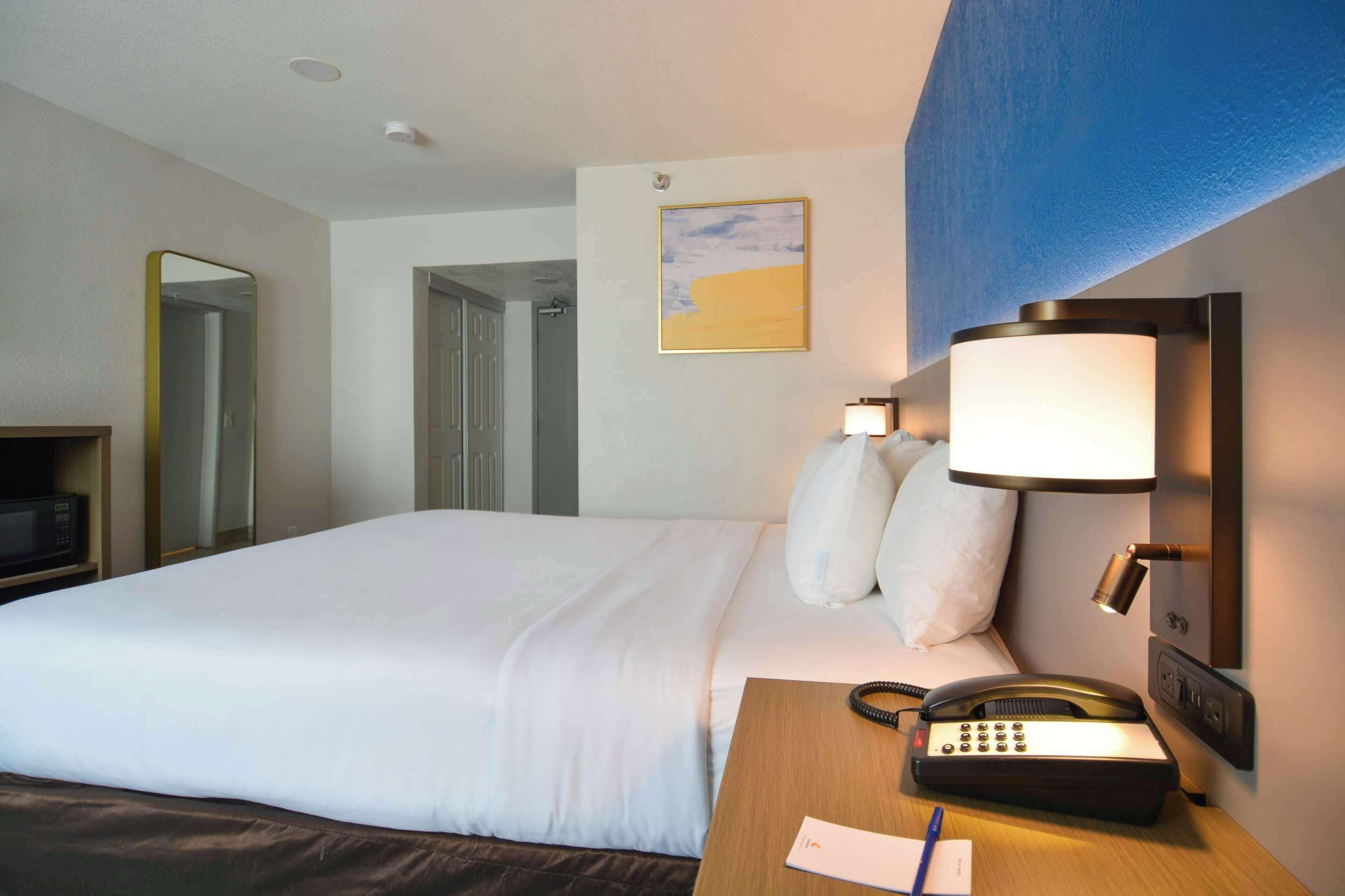 A hotel room with a large bed, a bedside telephone, a lamp, a mirror, and a painting on the wall.