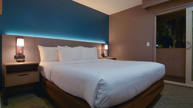 A hotel room with a large bed, two bedside tables with lamps, a nightstand, and a view of the outdoors through a sliding door.