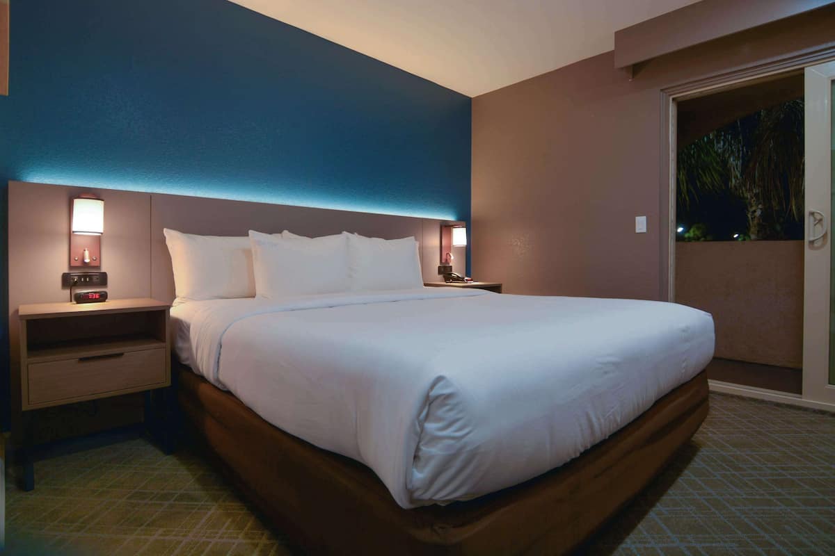 A hotel room with a large bed, two bedside tables with lamps, a nightstand, and a view of the outdoors through a sliding door.