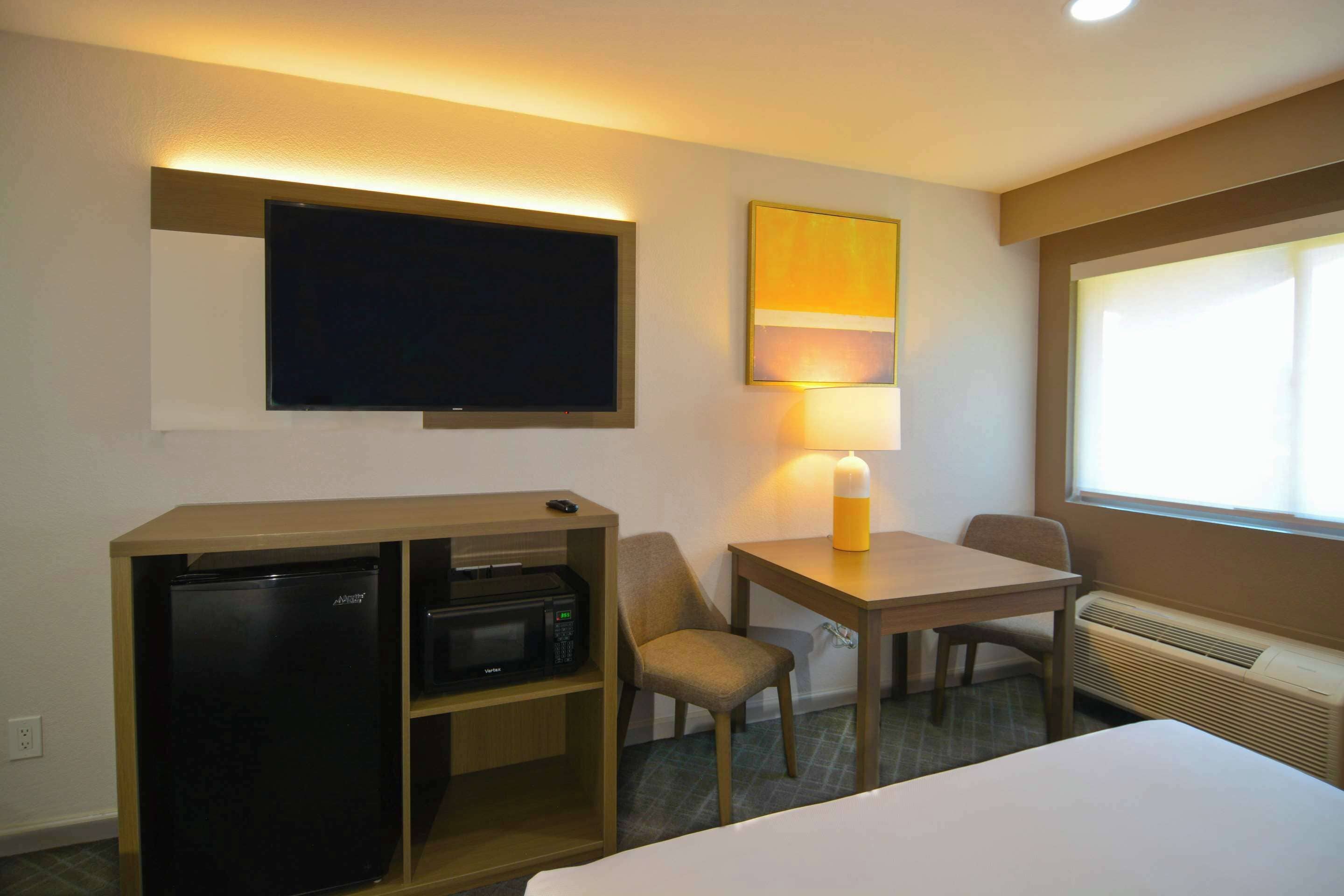 A hotel room with a bed, a desk with a chair, a small refrigerator, a microwave, a TV, a lamp, and a window with blinds.