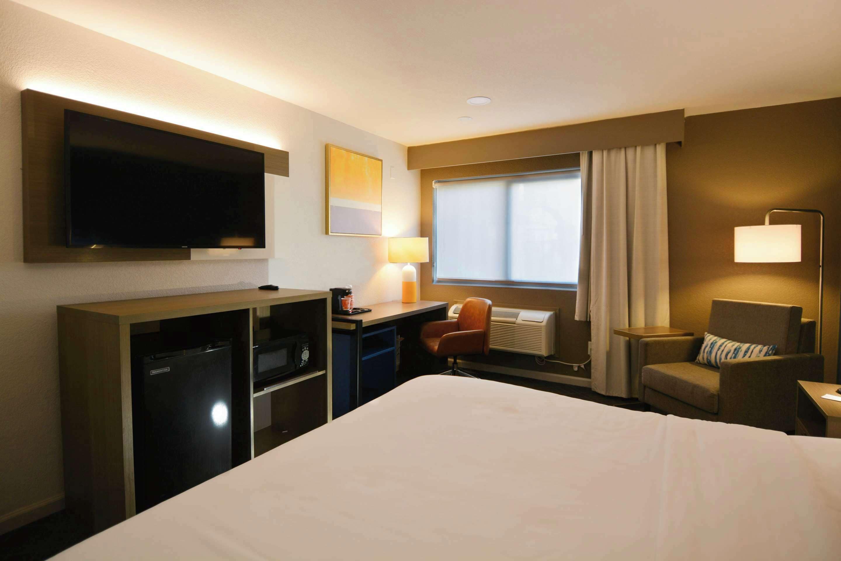 A hotel room with a large bed, a desk with a chair, a television, a lamp, and a window with curtains.