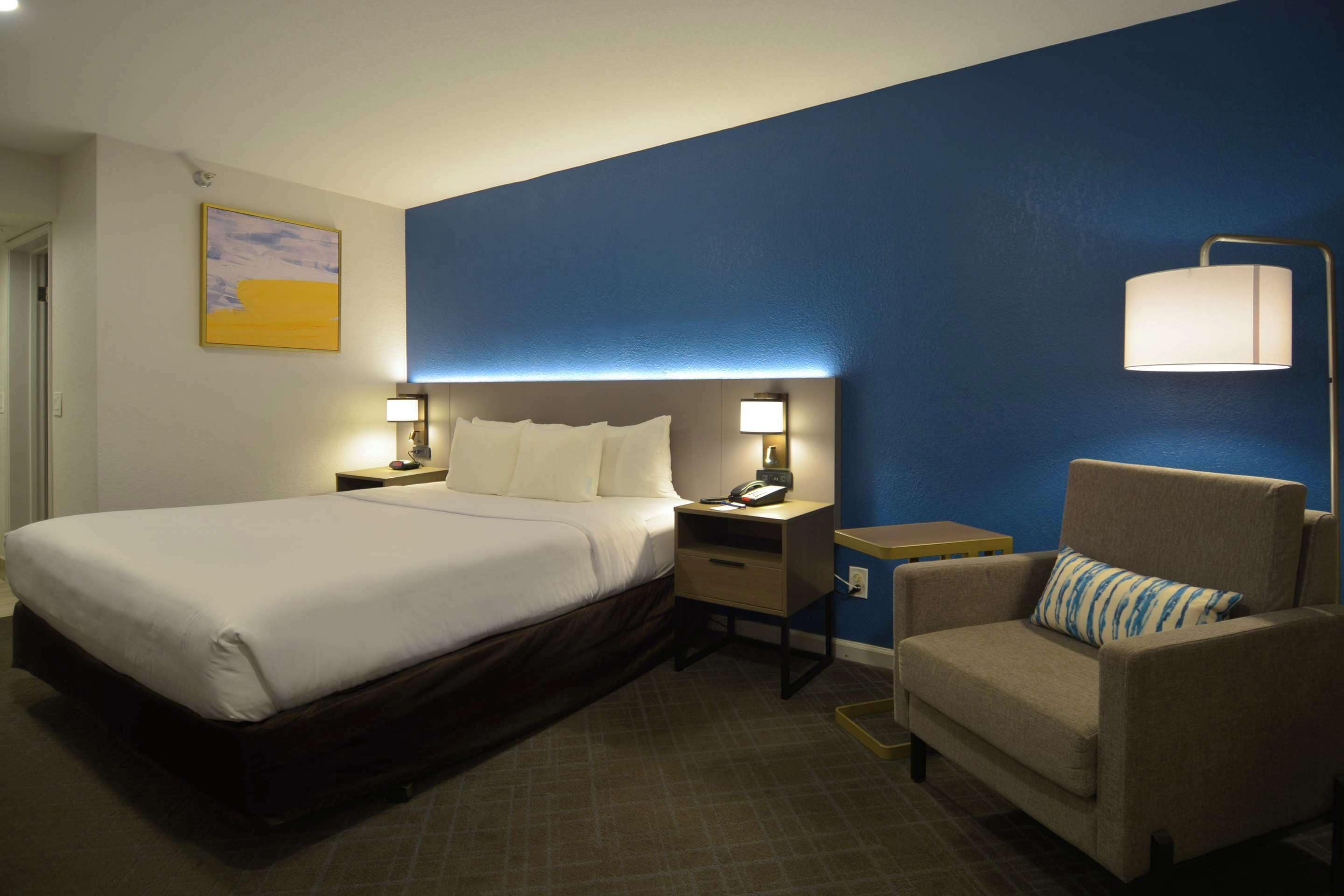 A hotel room with a large bed, bedside tables with lamps, a chair, and a blue accent wall.