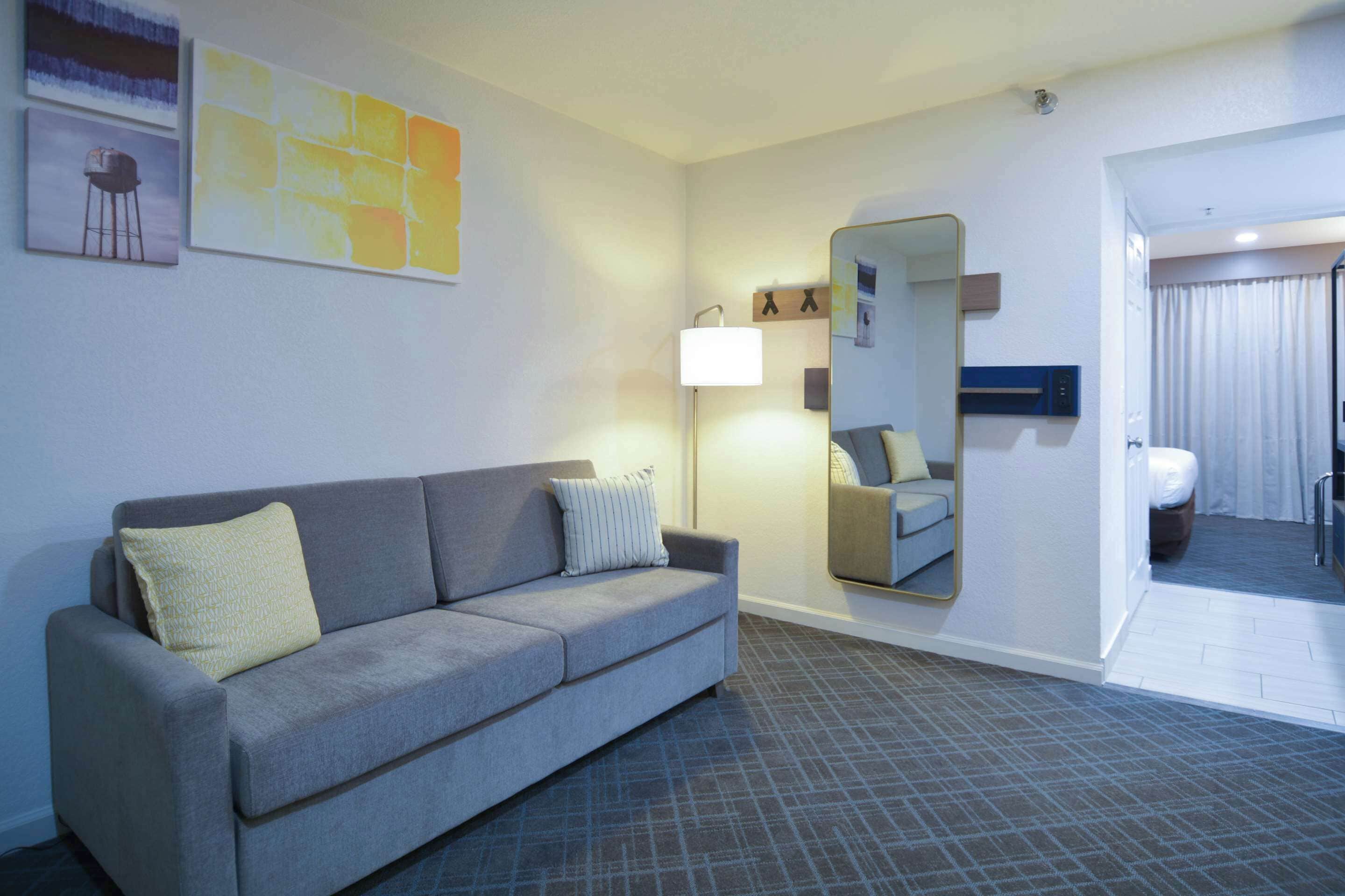 A modern hotel room with a grey sofa, a large mirror, a standing lamp, and a view into the bathroom.