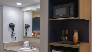 A hotel bathroom with a sink, mirror, hairdryer, microwave, coffee maker, and a refrigerator.