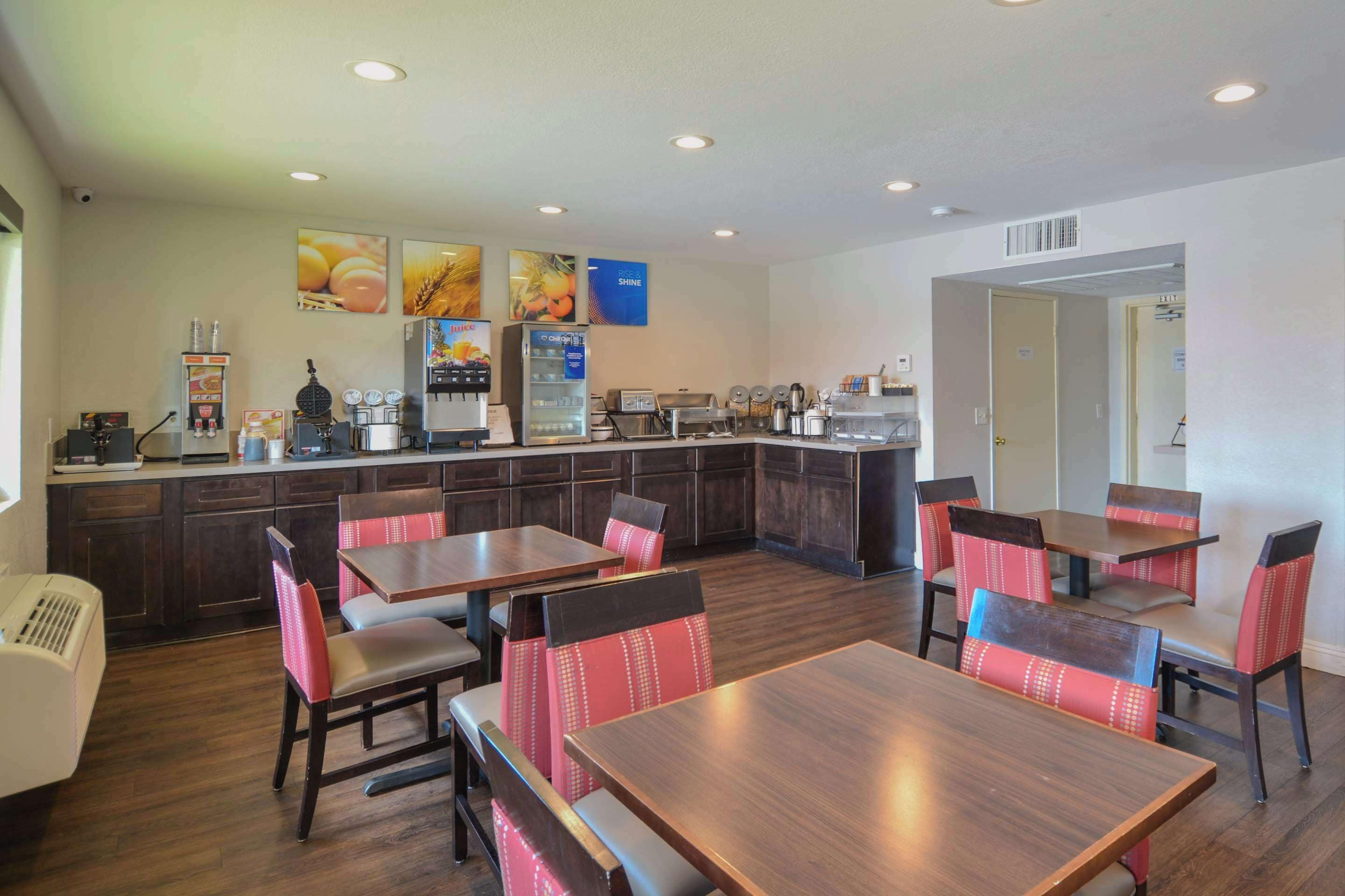A restaurant with wooden tables and red upholstered chairs, a kitchen area with appliances, and a wall with fruit and grain images.