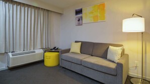 A modern hotel room with a grey sofa, a yellow ottoman, a standing lamp, and a radiator.