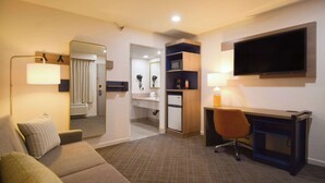 A hotel room with a sofa, a desk, a TV, a mirror, a bathroom with a sink and a hairdryer, and a shelf with a lamp.