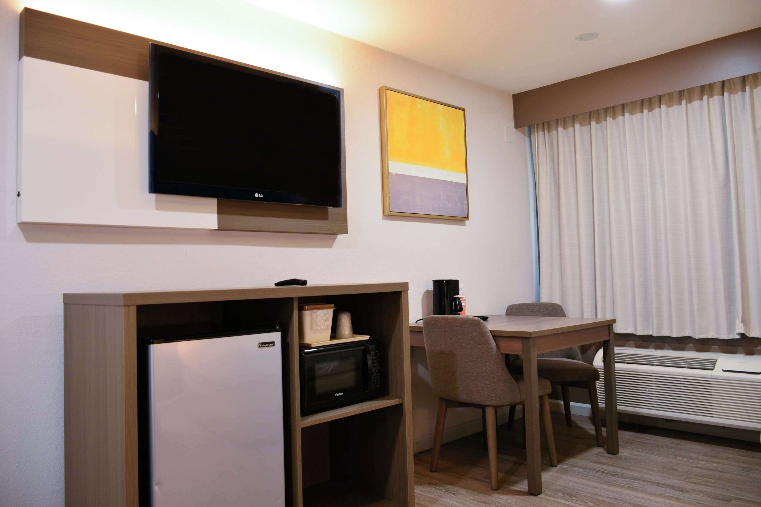 A hotel room with a flat-screen TV mounted on the wall, a desk with a chair, a microwave, and a refrigerator.