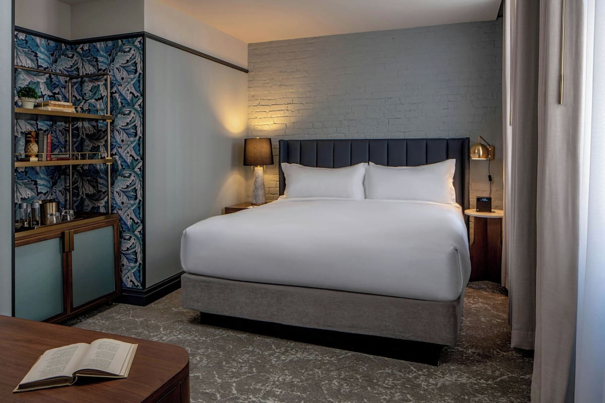 Habitación de hotel con una cama grande, una mesita de noche con lámpara, una estantería y una pequeña mesa con un libro.