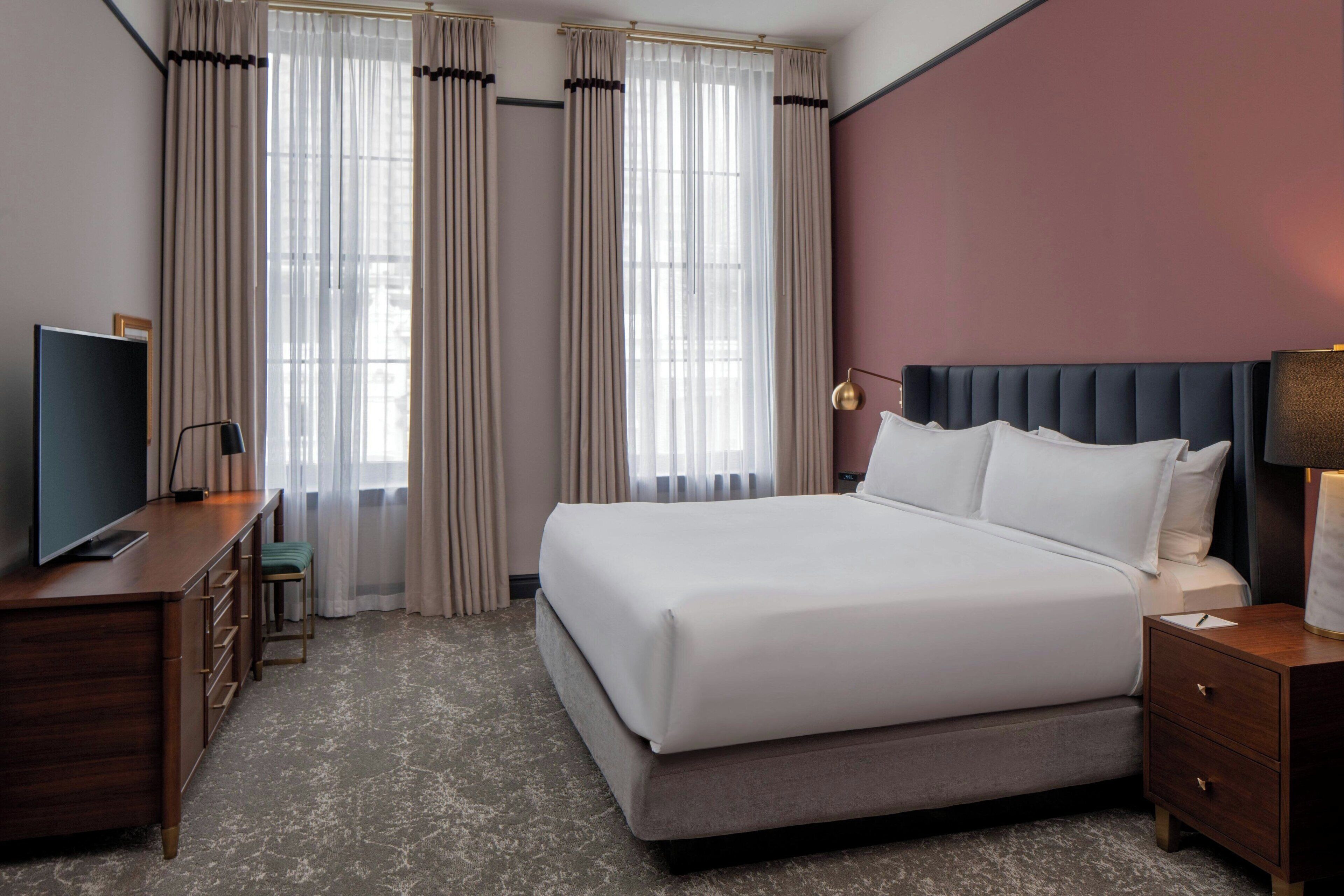 Quarto de hotel com uma cama, uma escrivaninha com televisão, um abajur e cortinas.