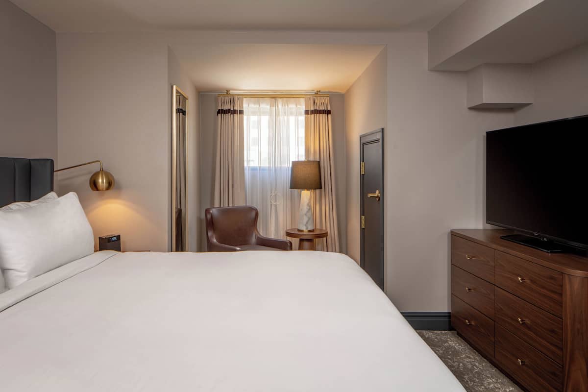 Una habitación de hotel con cama, una silla, televisión, lámpara y armario.
