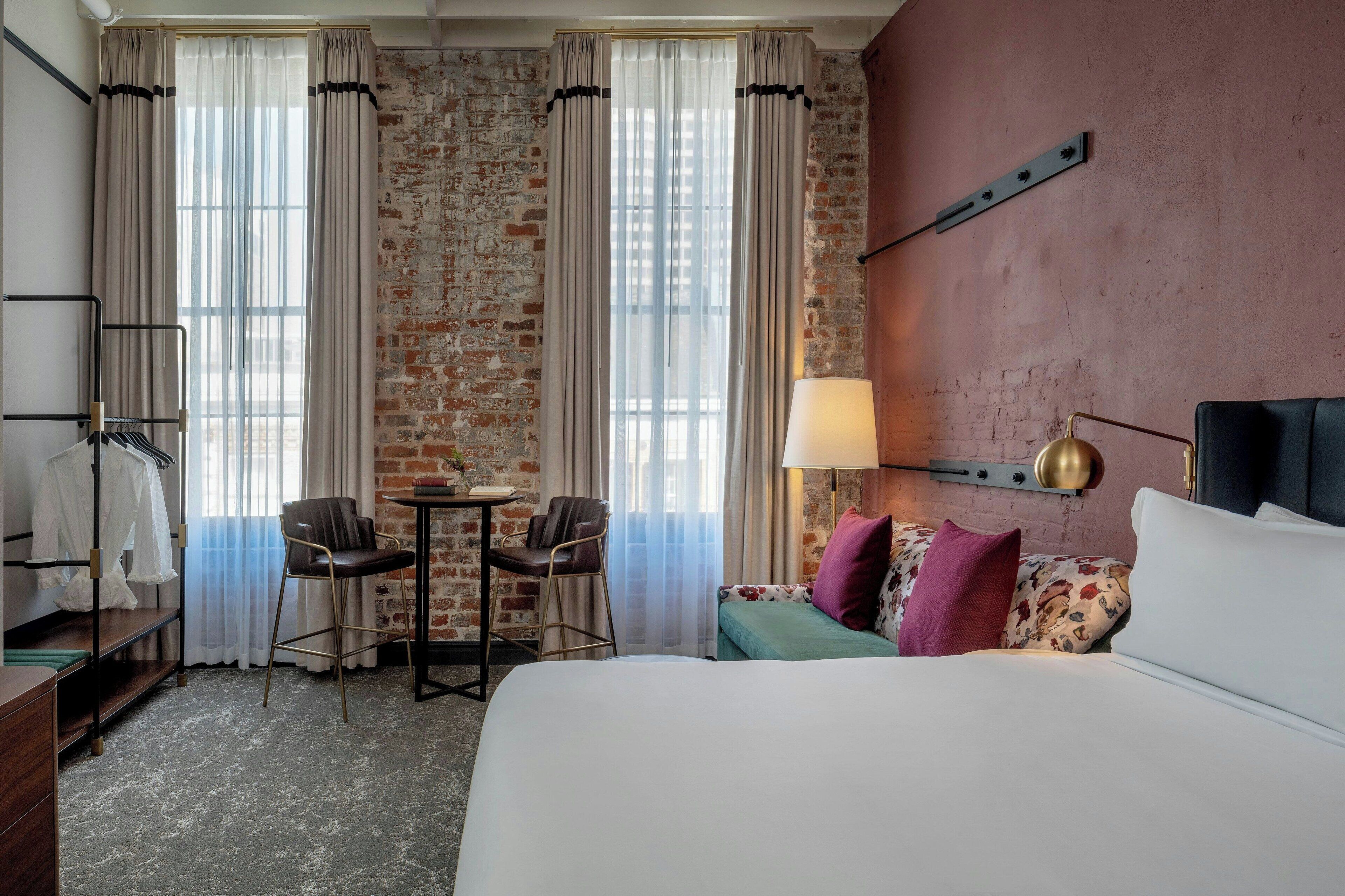 A hotel room with a bed, a desk, a chair, a small table, and a lamp.