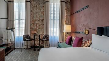 A hotel room with a bed, a desk, a chair, a small table, and a lamp.