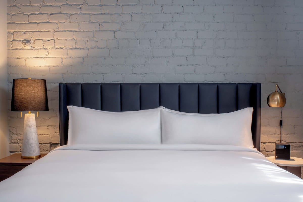 Una cama con cabecera oscura, una lámpara de noche, un reloj y una pared con diseño de ladrillo.