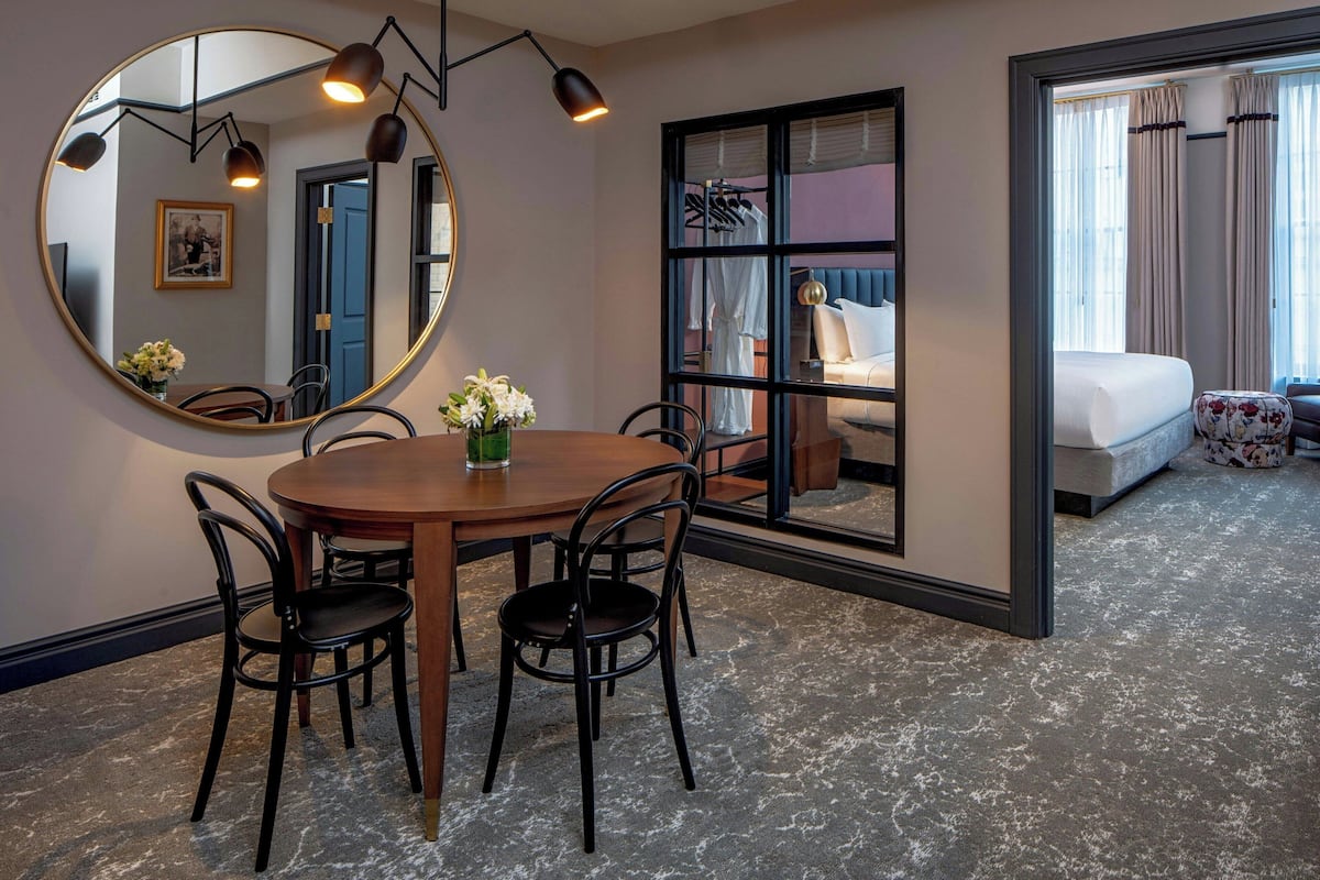 Habitación de hotel con una mesa de comedor redonda, cuatro sillas, un espejo grande, una ventana con cortinas, una cama y una pequeña mesa con un jarrón de flores.