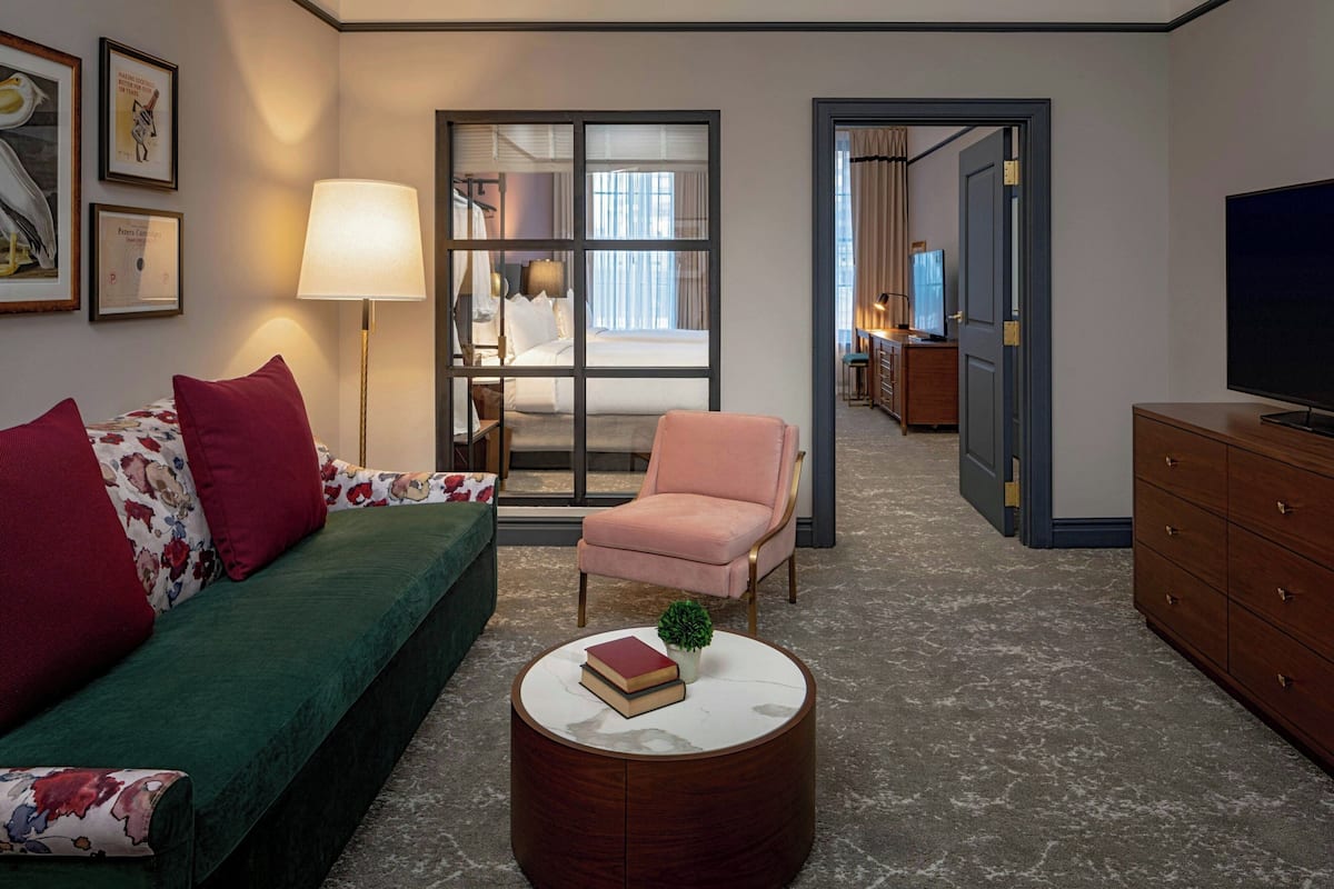 Habitación de hotel con sofá verde, sillón rosa, una mesita de centro redonda con libros, televisor y vistas a un dormitorio a través de un ventanal.