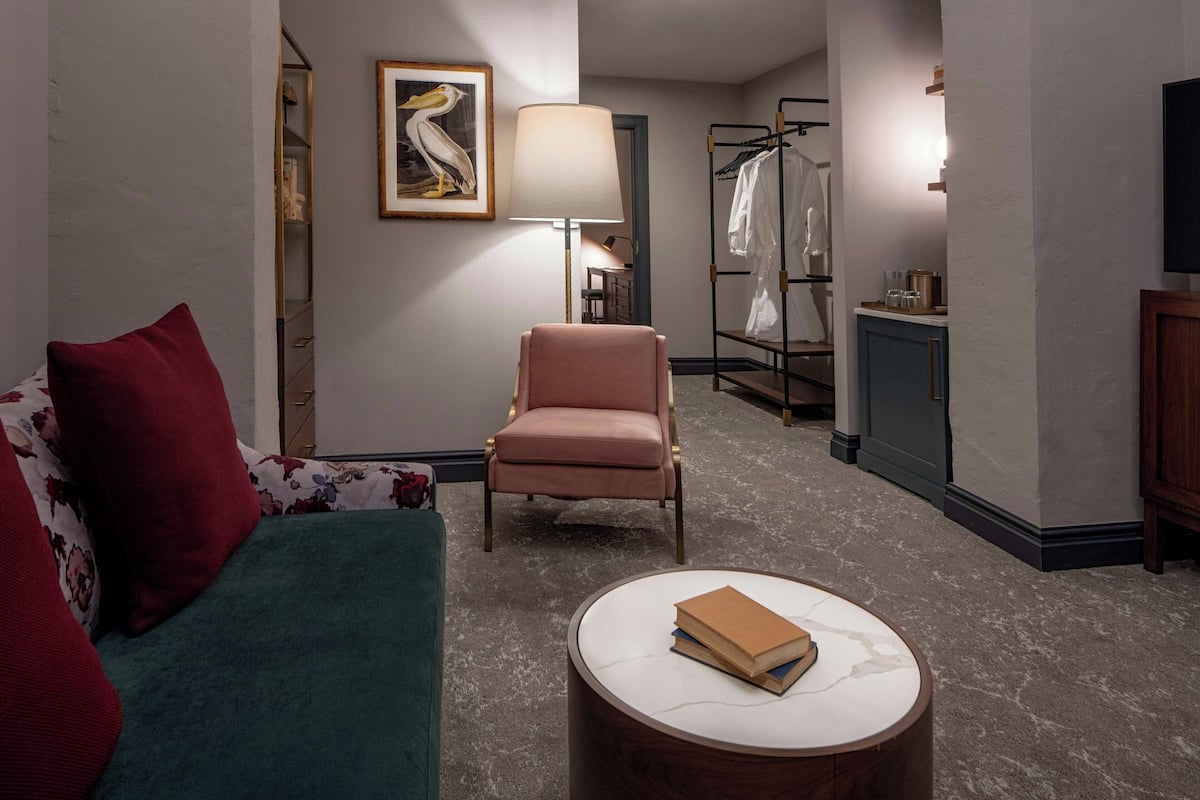 Habitación de hotel con sofá verde, sillón rosa, una mesita de centro redonda con libros, una lámpara, perchero y televisor.