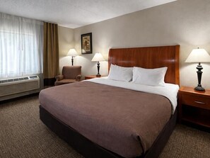 A hotel room with a large bed, two bedside tables with lamps, a chair, a window with curtains, and a wall-mounted picture.