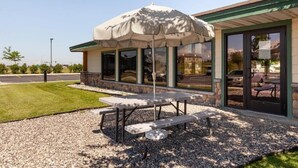 An outdoor patio with a table and benches, a large umbrella, and a building with large windows and a stone facade.