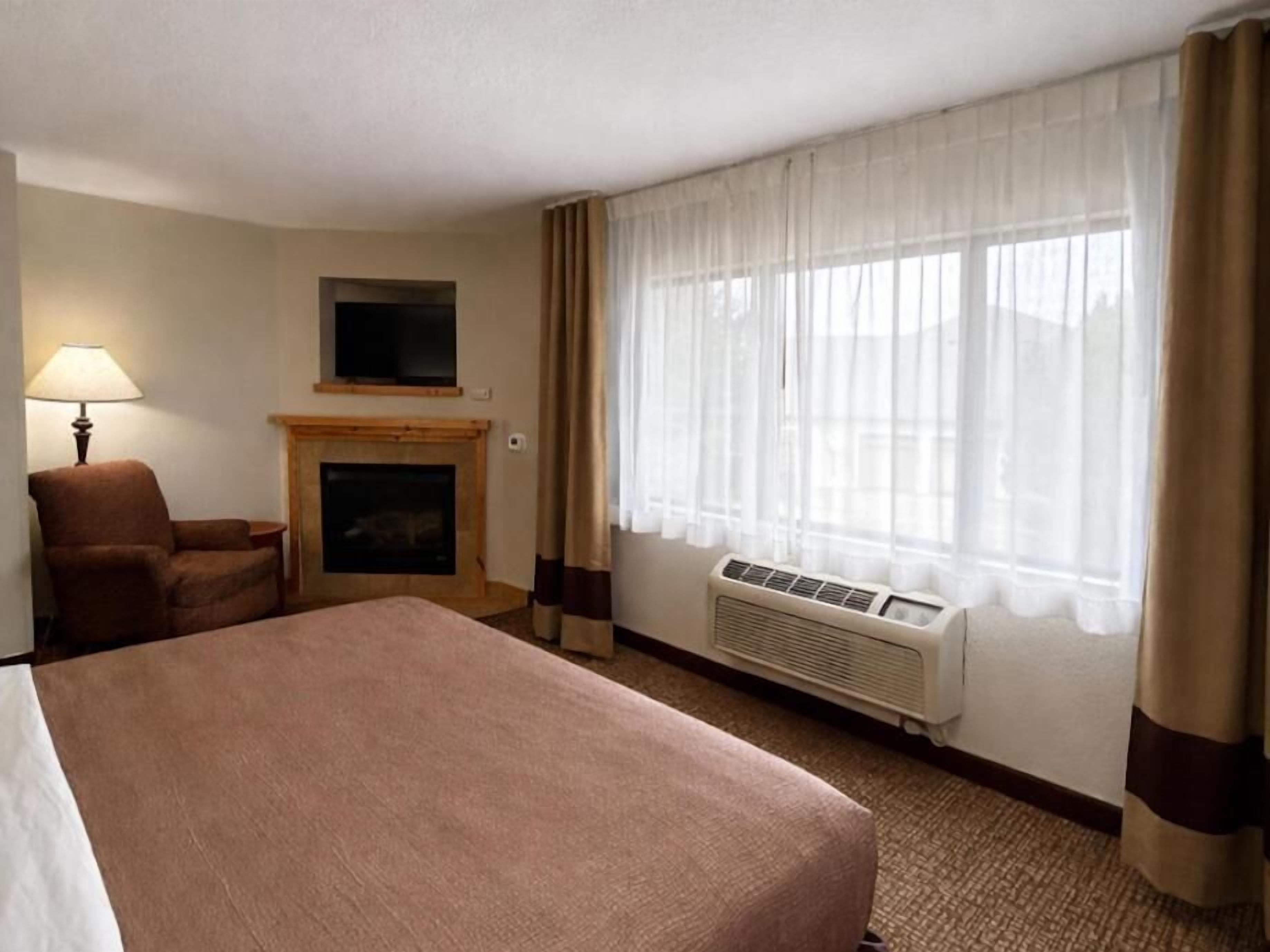 Une chambre d’hôtel avec un lit, une chaise, une cheminée, une télévision et une fenêtre avec des rideaux.