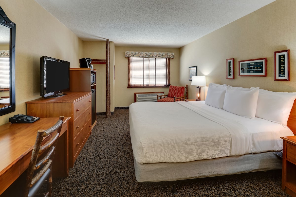 A hotel room with a bed, a desk with a computer, a television, a window with blinds, and a wall-mounted air conditioner.