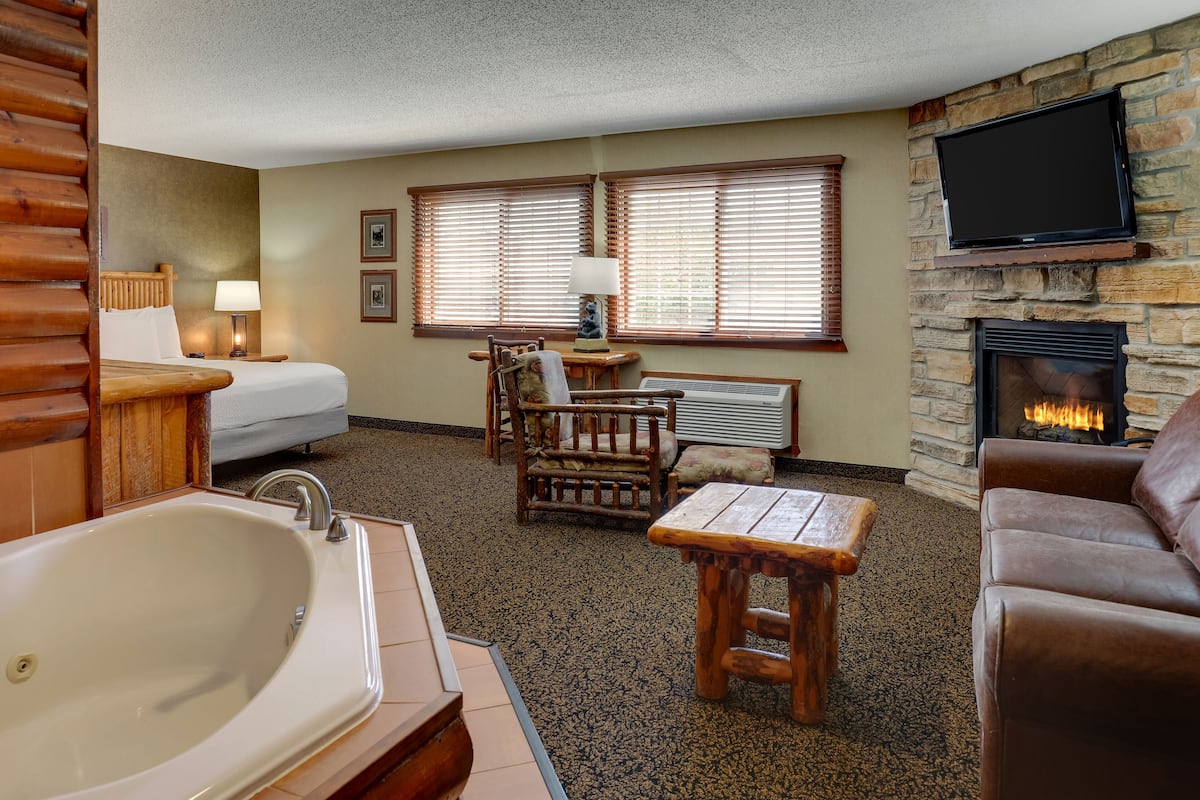 A room with a large bathtub, a fireplace, a television mounted on a stone wall, and a bed with a wooden headboard.