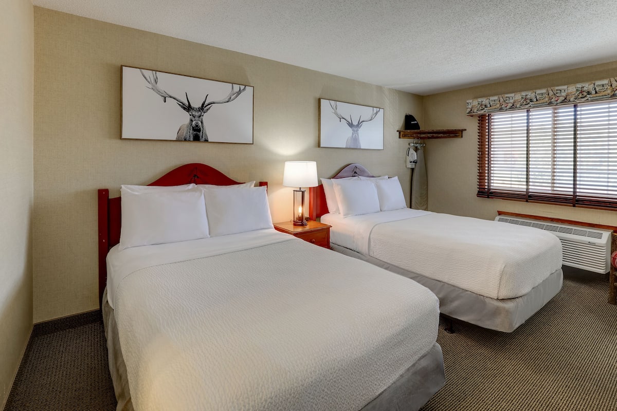 A hotel room with two beds, a nightstand with a lamp, a window with blinds, and two wall-mounted deer artwork.