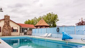 A swimming pool with lounge chairs and a covered area, surrounded by a fence and a building with a stone chimney.