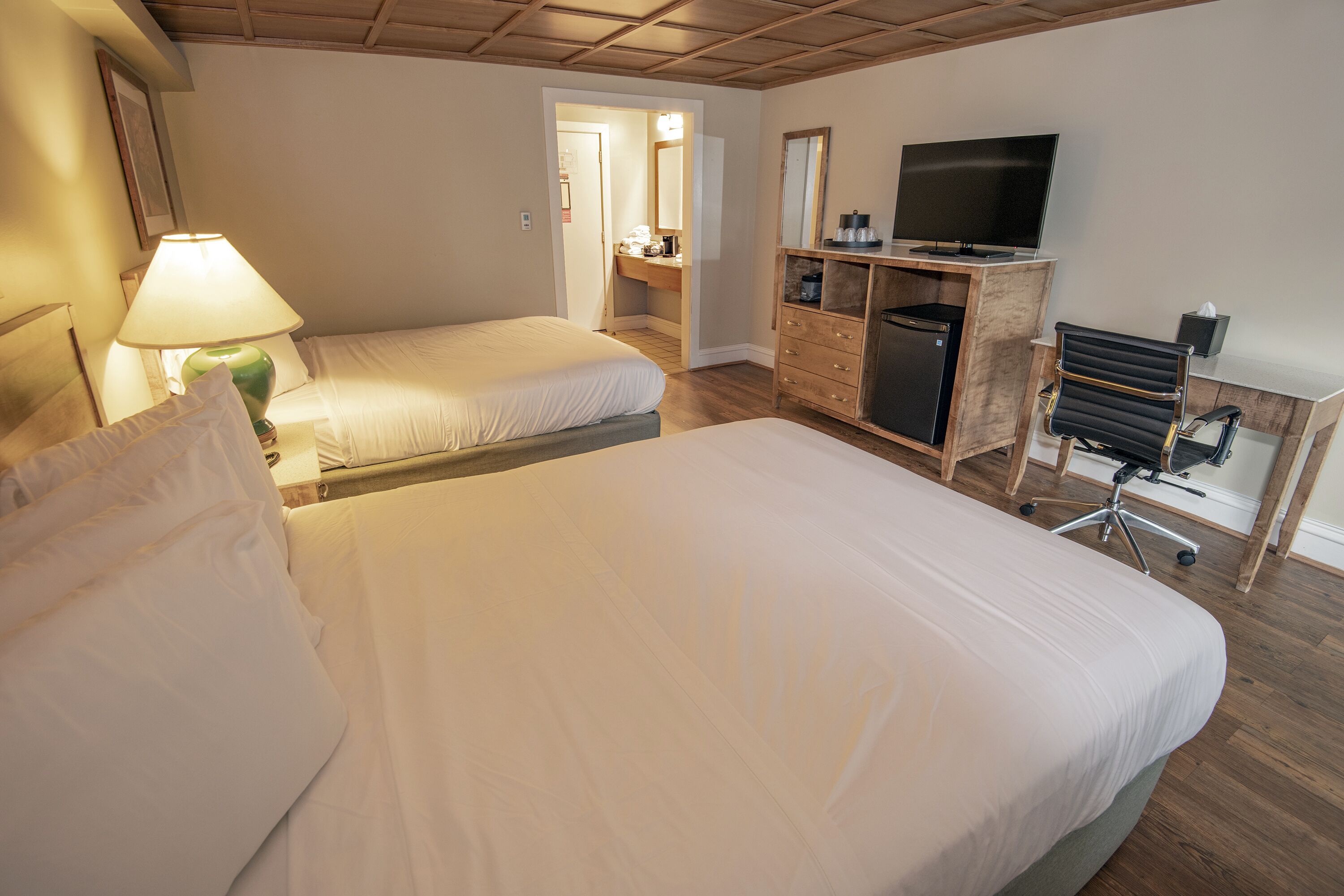 Habitación de hotel con dos camas, un escritorio con computadora, un televisor y un baño visible a través de una puerta abierta.