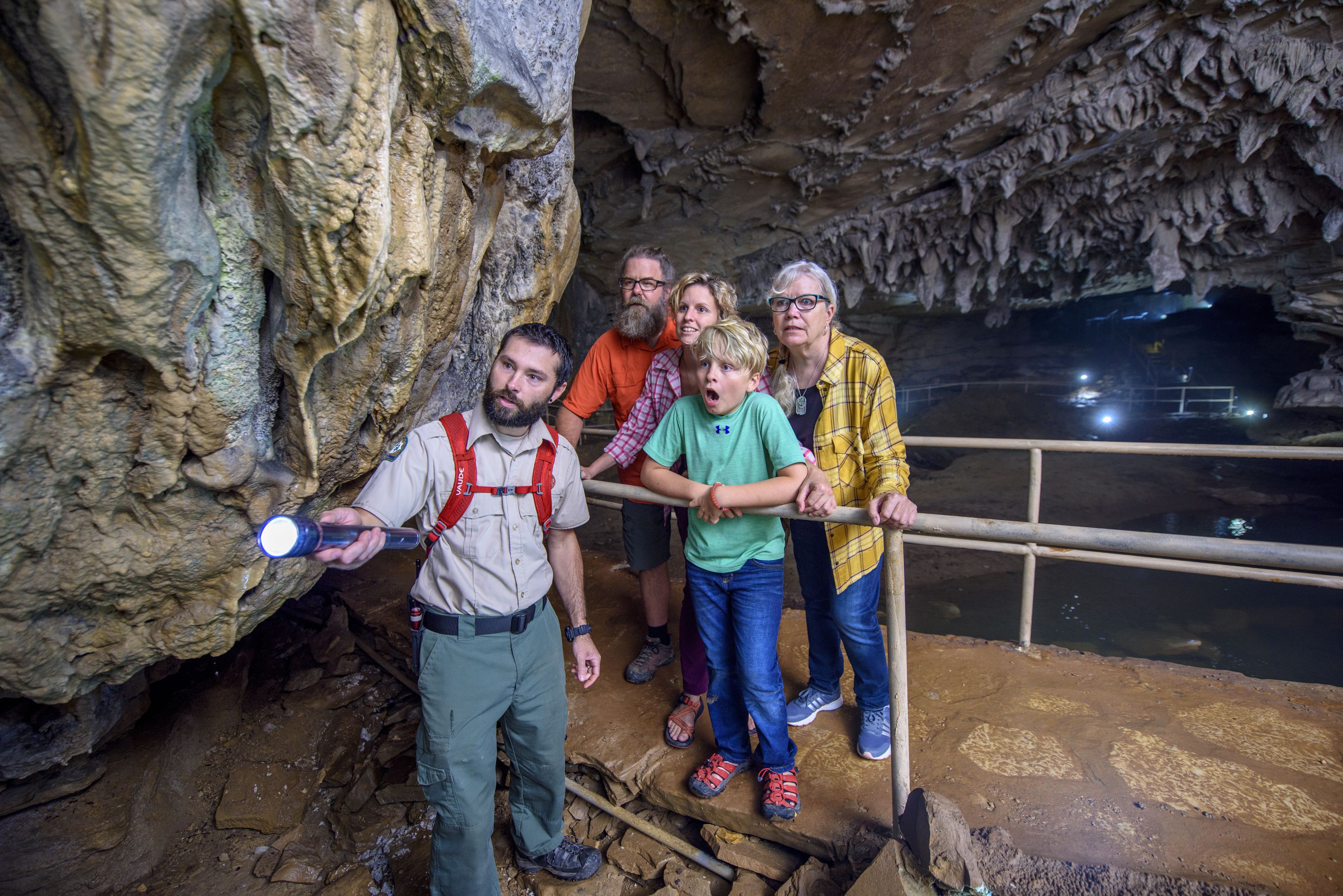 A group of people inside a cave with rocky walls and a railing overlooking a body of water.