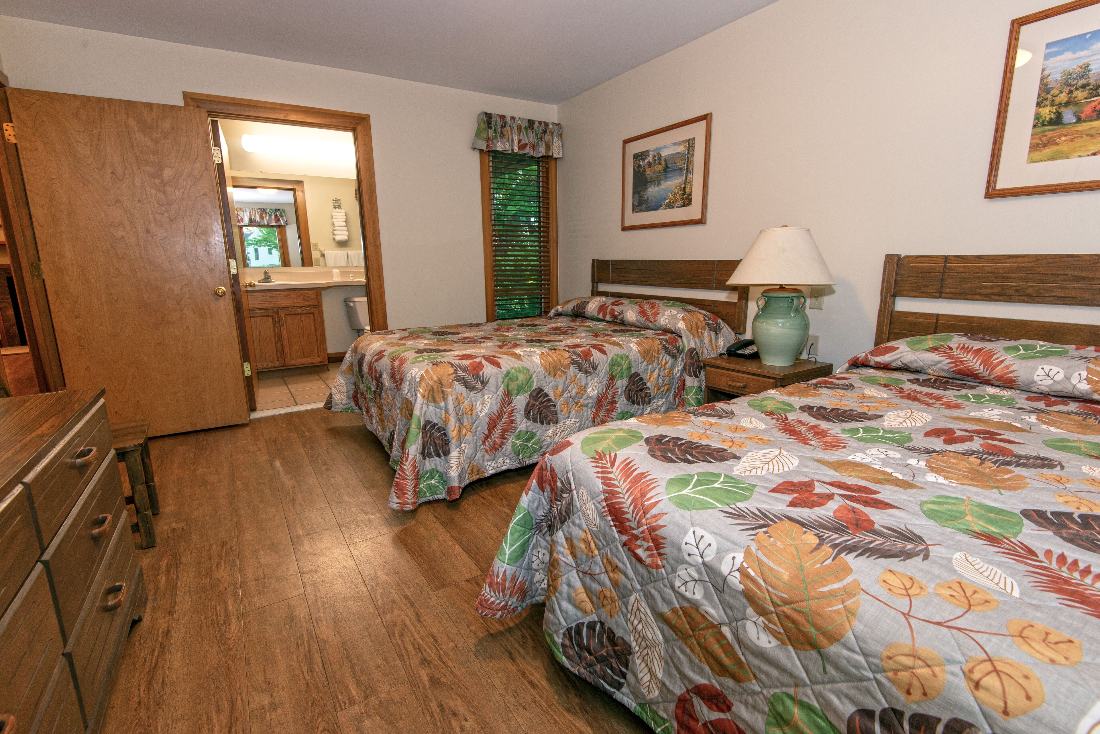 Un dormitorio con dos camas, piso de madera, una mesita de noche con lámpara, un aparador y una puerta que da a otra habitación.