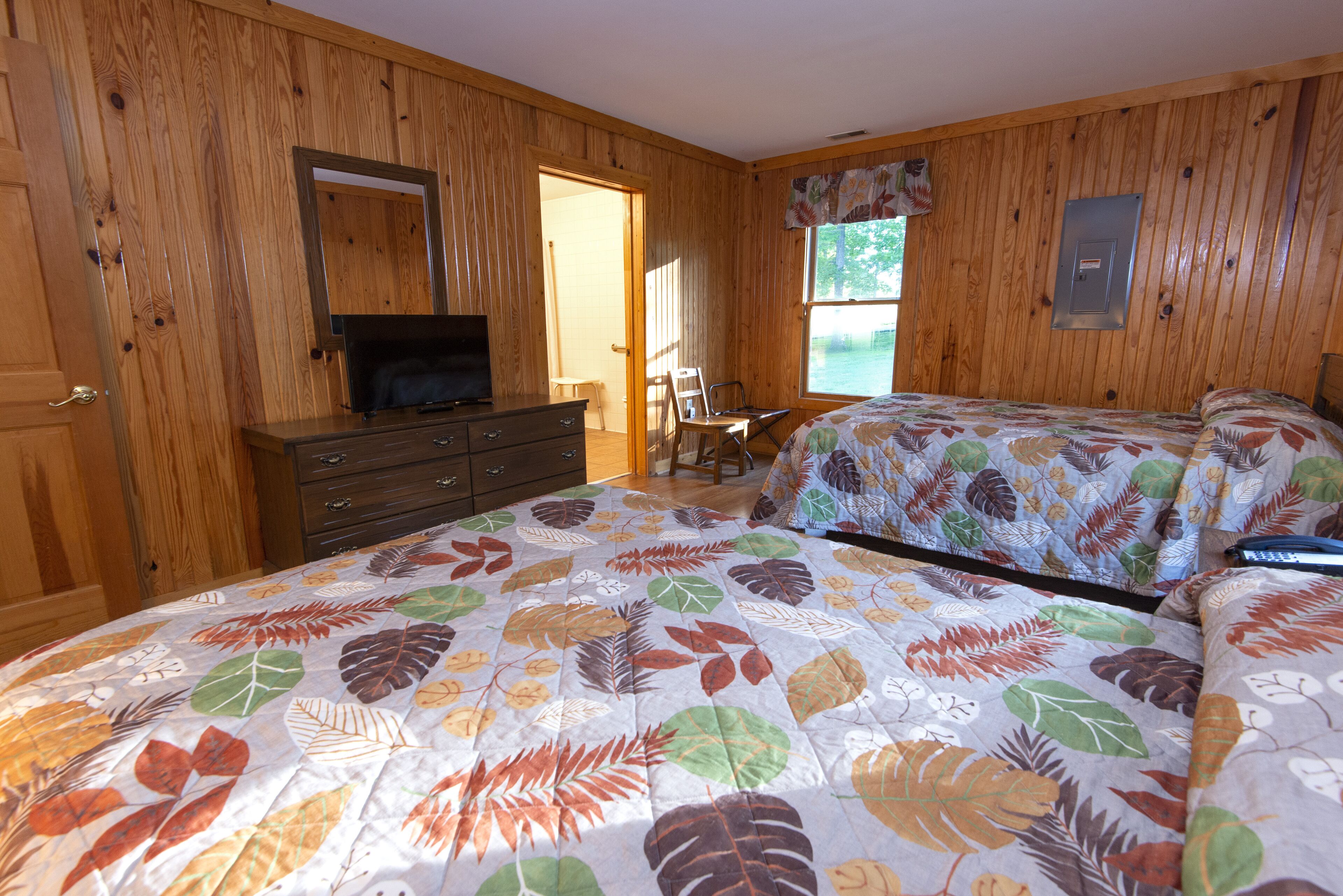 A bedroom with wooden walls, a bed with a leaf-patterned bedspread, a dresser, a chair, and a television.