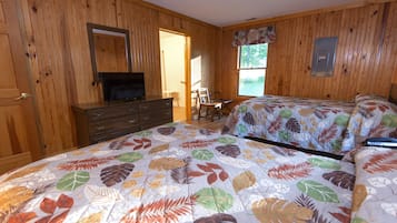 A bedroom with wooden walls, a bed with a leaf-patterned bedspread, a dresser, a chair, and a television.