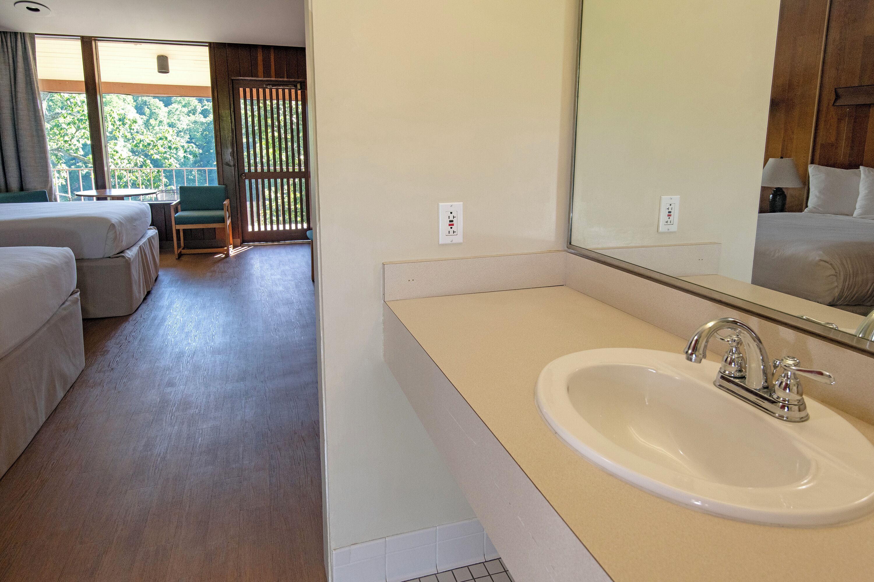 Habitación de hotel con una cama grande, una mesita, una silla, un baño con lavamanos y espejo, y vistas al exterior.