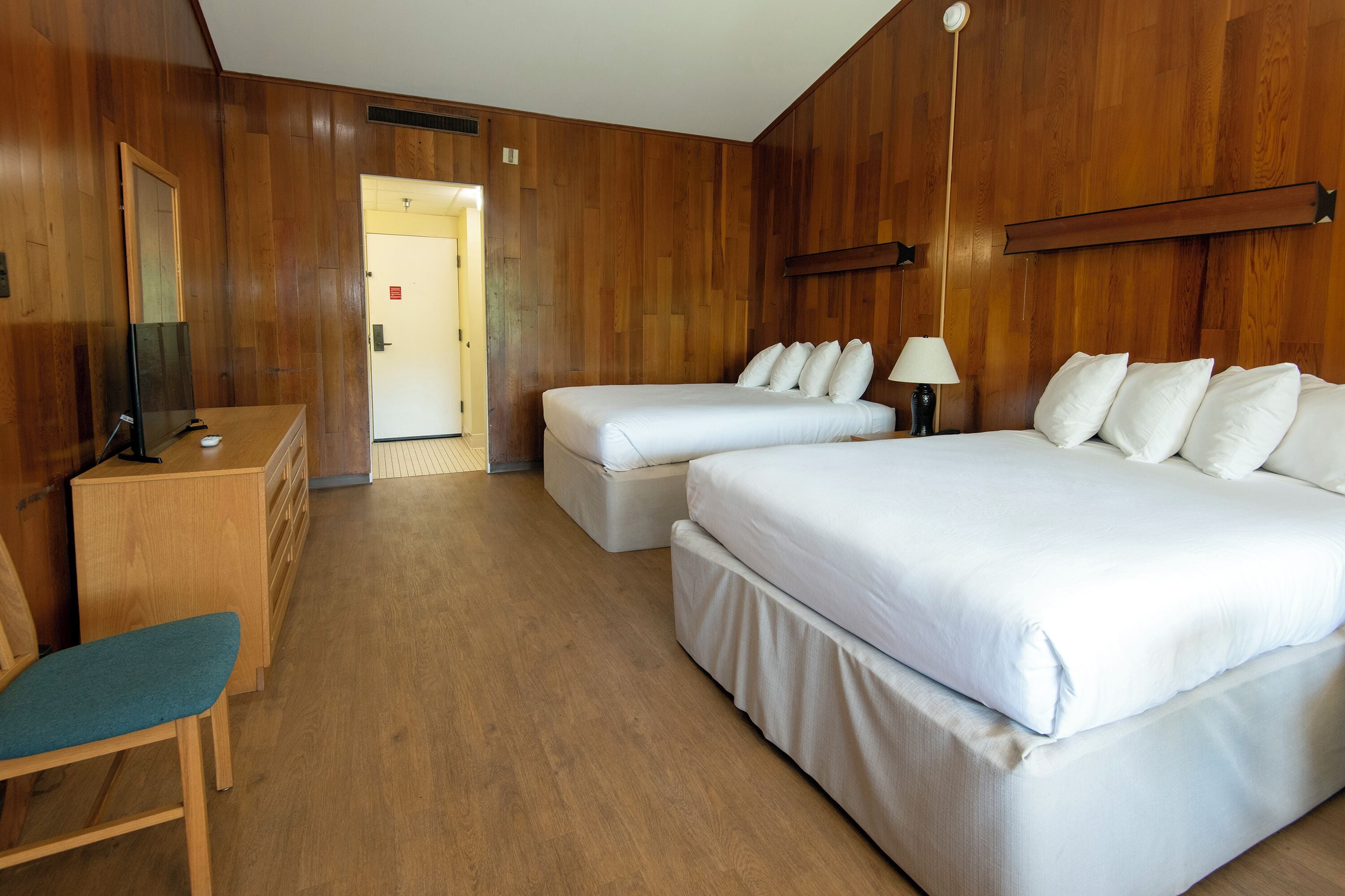 Quarto de hotel com duas camas, uma escrivaninha e uma cadeira, e paredes de madeira.