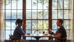 Deux personnes sont assises à une table en bois, devant de grandes fenêtres qui donnent sur un lac.