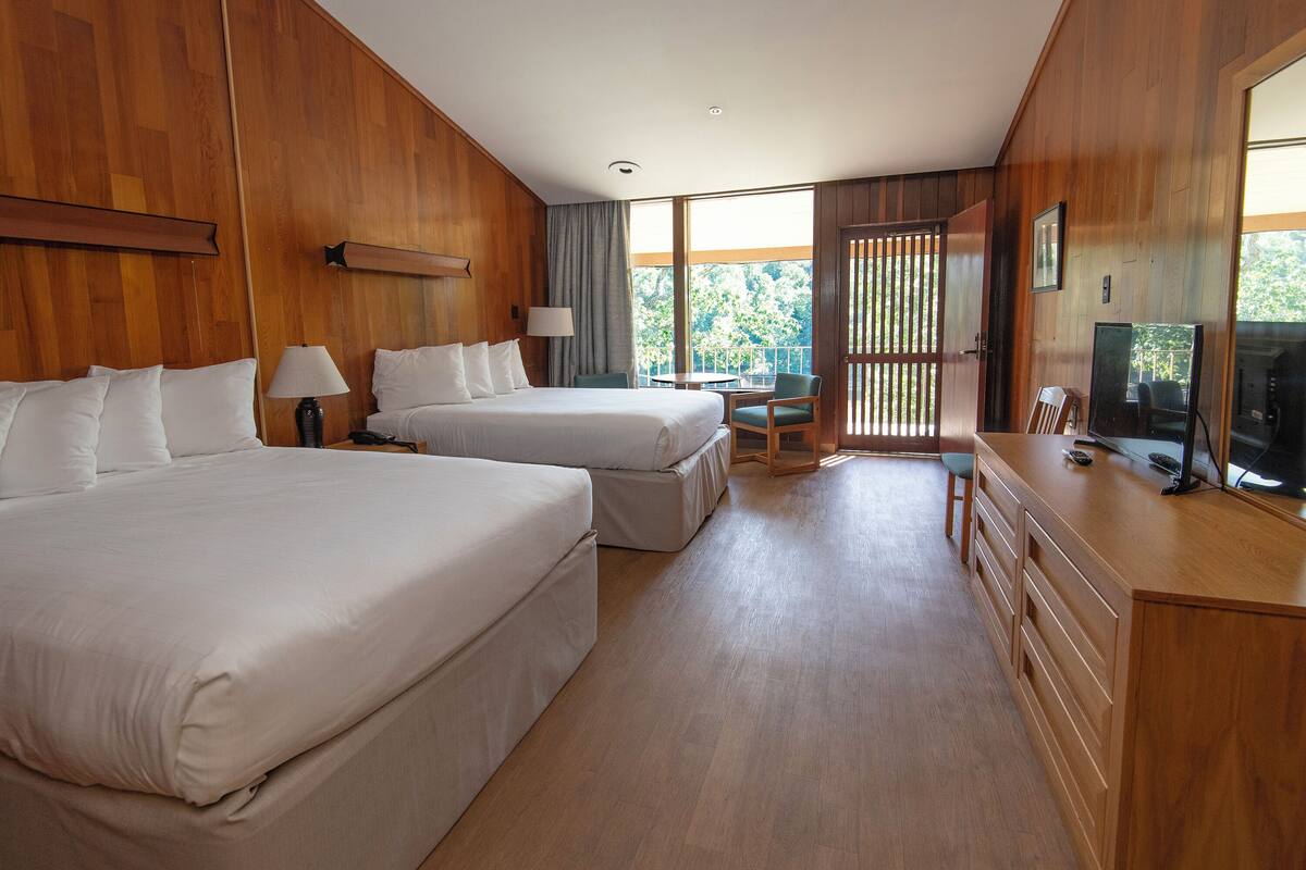 A hotel room with two beds, a desk, a chair, a TV, and a large window with a view of trees.