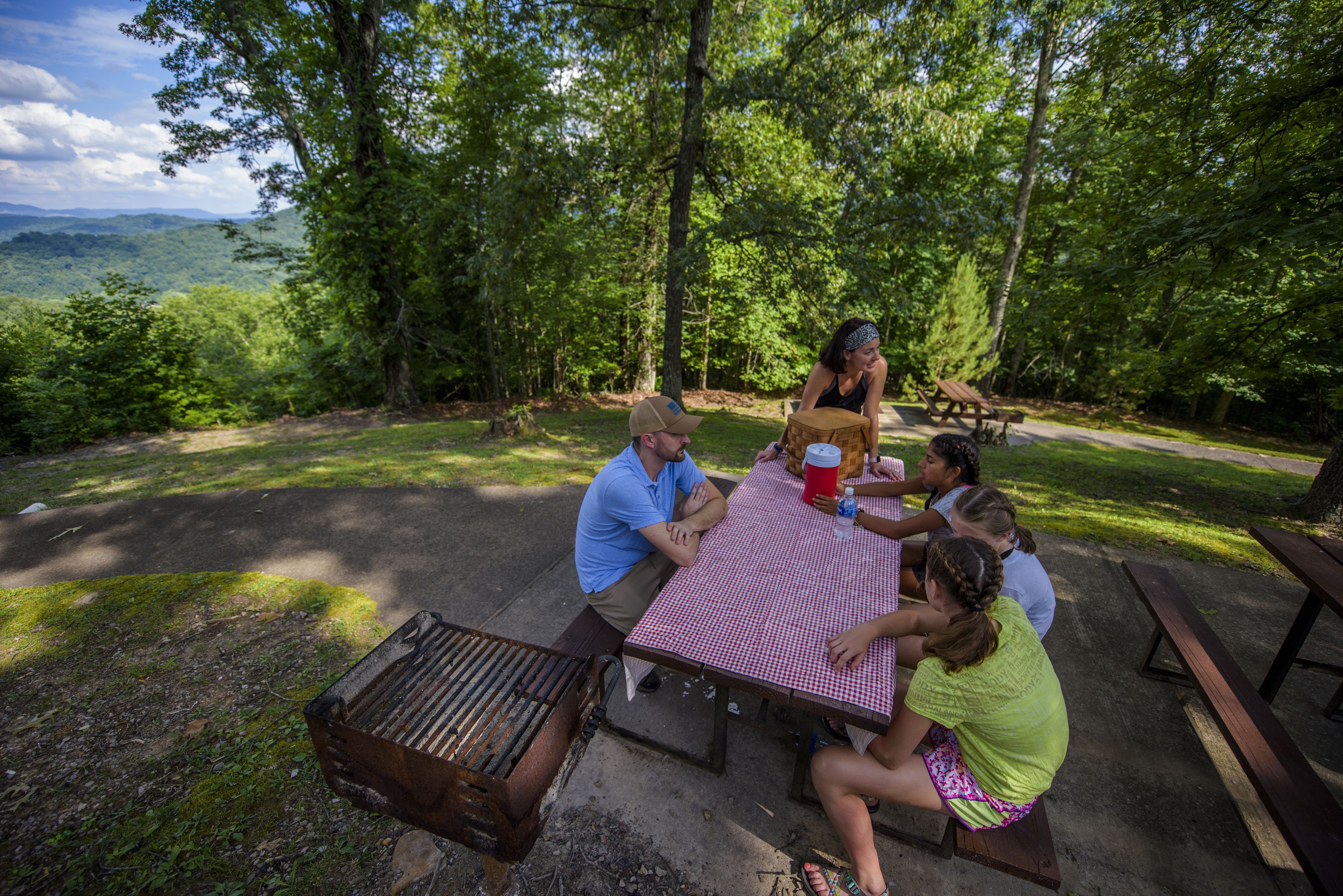 Eine Gruppe von Menschen sitzt an einem Picknicktisch im Freien mit Blick auf einen Wald und Berge im Hintergrund.
